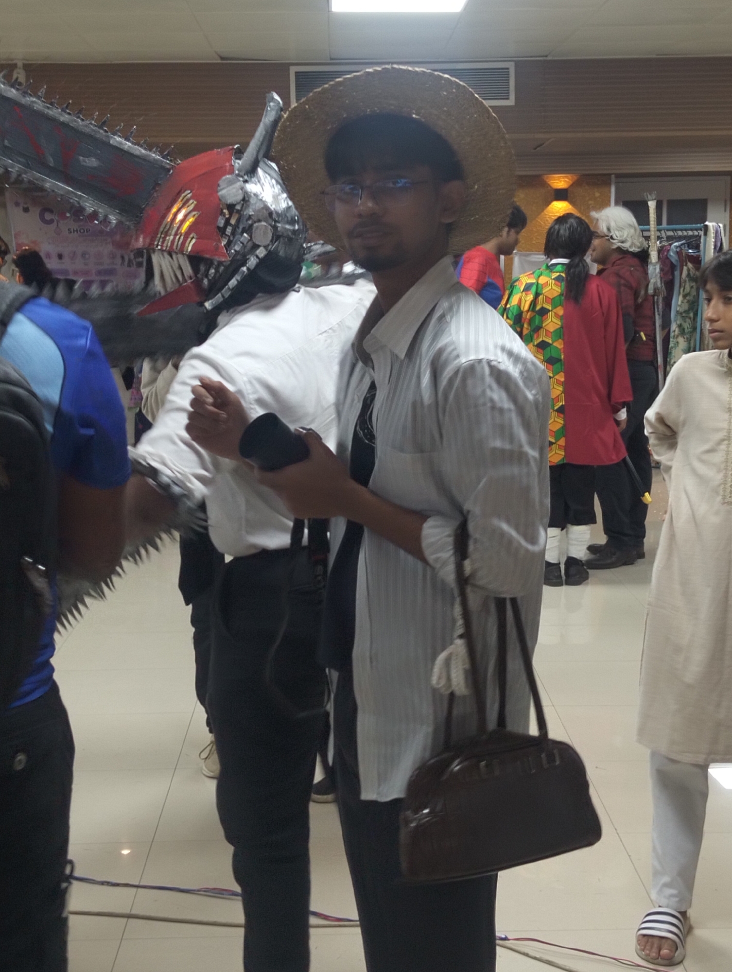 The image depicts a gathering of individuals in what appears to be an indoor event, possibly a convention or a cultural exhibition. The setting is illuminated by overhead lighting, and the floor is tiled. In the foreground, a man wearing a straw hat and glasses is partially visible, holding a dark cylindrical object in his right hand. He is dressed in a light-colored, striped shirt and dark pants. To his left, another individual is seen from the shoulder down, wearing a blue and black outfit and a large, elaborate headpiece that resembles a chainsaw.
In the background, several other people are present. One person is wearing a red and green patterned garment and dark pants. Another individual, to the right, is dressed in a light-colored traditional outfit. Various items, including clothing racks and banners, are visible, suggesting a public event or marketplace. The overall atmosphere appears casual and interactive, with people congregating and observing. The presence of cosplay elements, like the chainsaw headpiece, points towards a fan convention or similar event. The specific location in the city of Dhaka, Bangladesh, cannot be determined from the visual information provided.