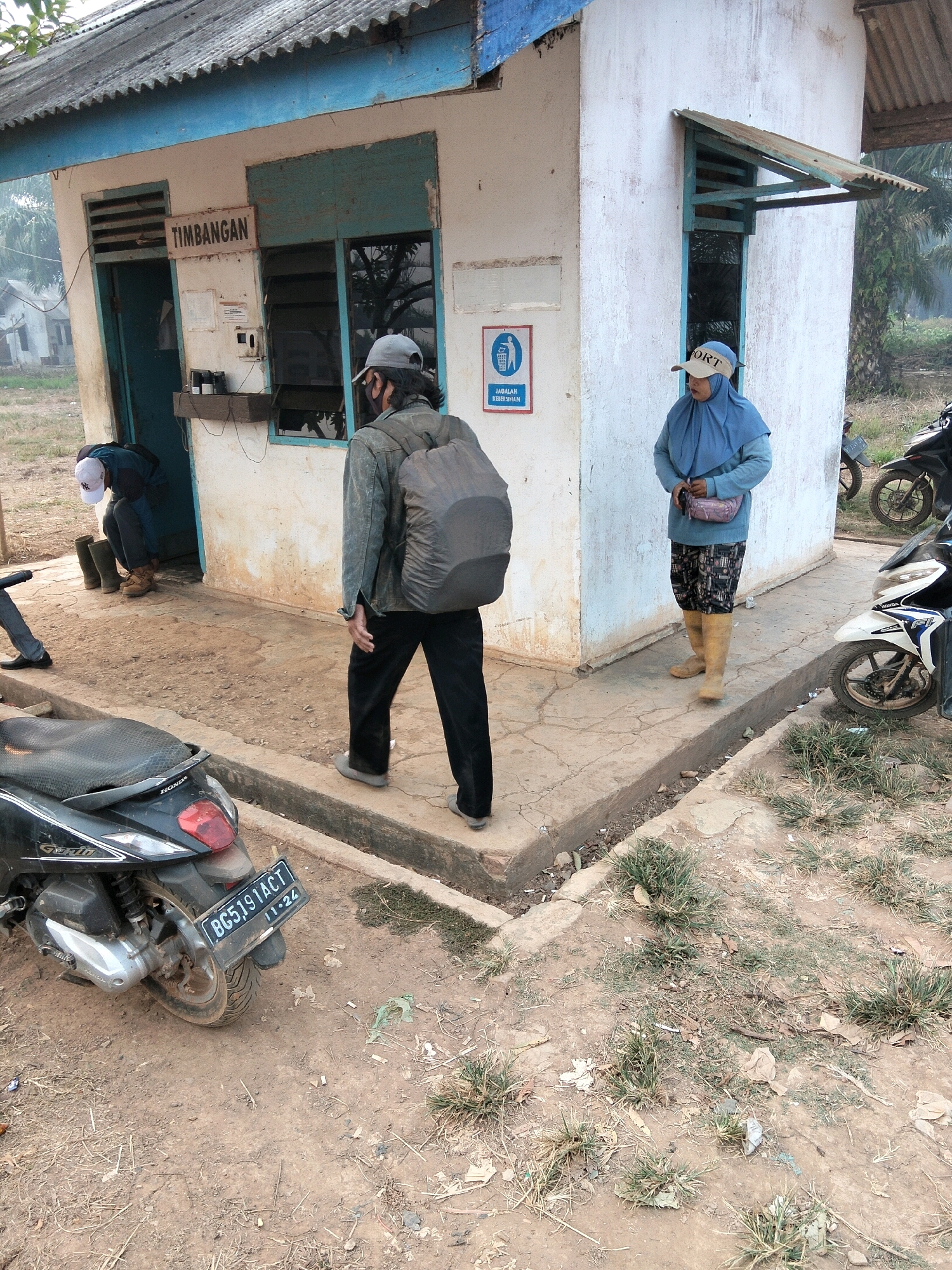 The image depicts an outdoor scene in what appears to be Teluk Tenggirik, Indonesia, likely near a weigh station or small administrative building. A white, two-story building with a corrugated metal roof and blue trim is the central structure. A sign above a door reads "TIMBANGAN," which translates to "Weighing Station."
In the foreground, a black Honda scooter is parked on a dirt path. A license plate with the characters "BG5191ACT 11-24" is visible. To the right of the building, another motorcycle, partially visible, is also present.
Several individuals are present. A person wearing a gray jacket, dark pants, and a backpack is walking away from the building. Another person, dressed in a blue headscarf and matching long-sleeved shirt, patterned trousers, and tall yellow boots, stands facing the building. A third individual, wearing a white cap and dark clothing, is bent over near the entrance of the building, possibly putting on or taking off boots.
The ground is a mix of dirt, gravel, and sparse vegetation. The overall impression is of a utilitarian, rural or semi-rural setting in Indonesia.