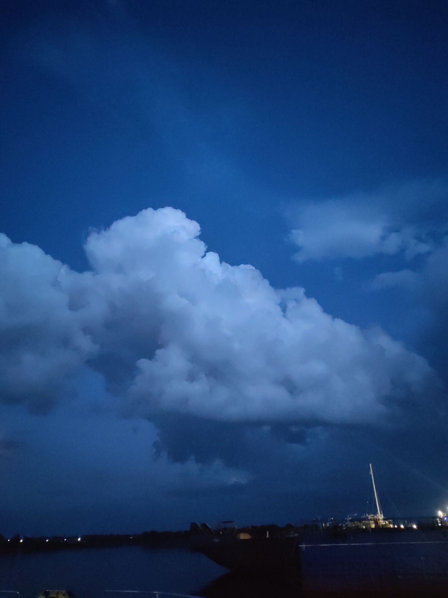 The image shows a dark blue sky with dramatic, white cumulus clouds. In the distance, a silhouette of a harbor is visible, with lights twinkling along the shoreline. A tall mast of a sailboat stands out against the darkening sky. The water in the foreground is calm and reflects the dim lights. The scene suggests it is either dusk or pre-dawn. The location is the city of Rybinsk, Russia.