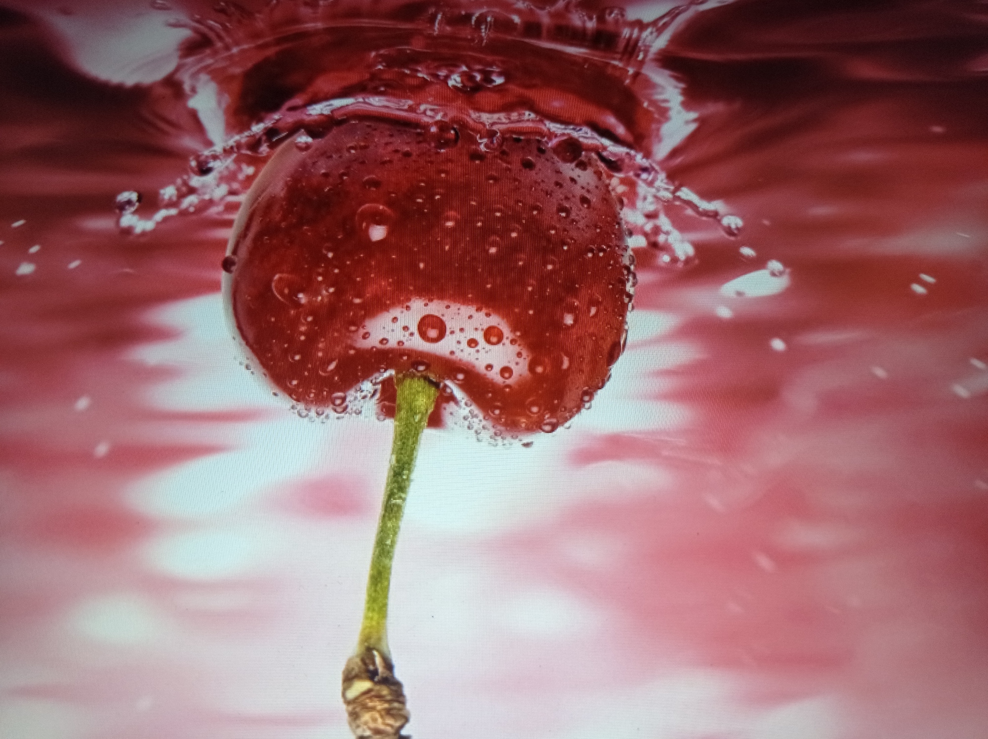 The image is a close-up, high-angle shot of a single, ripe red cherry with a stem, submerged in a deep red liquid, possibly juice or water, causing a splash. Water droplets are visible on the cherry's surface. The background is a blurry, gradient of pink and red hues. There is no discernible indication of the city of Mukachevo, Ukraine, or any urban environment present in this image.