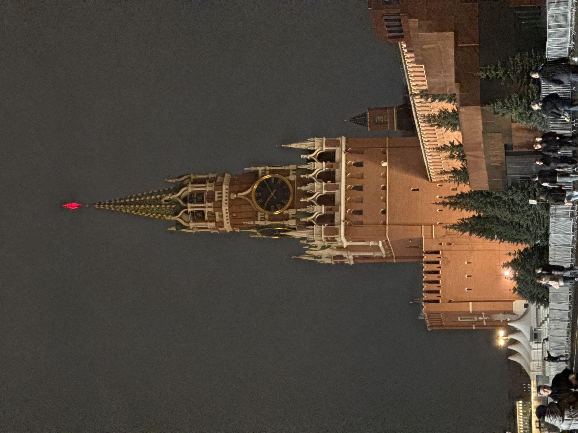 The image captures the Moscow Kremlin at night. The Spasskaya Tower dominates the scene with its clock face illuminated. A red star is visible at the top of the tower. Adjacent to the tower is a portion of the Kremlin wall, trees, and people walking. The environment is dark, indicating it is nighttime.