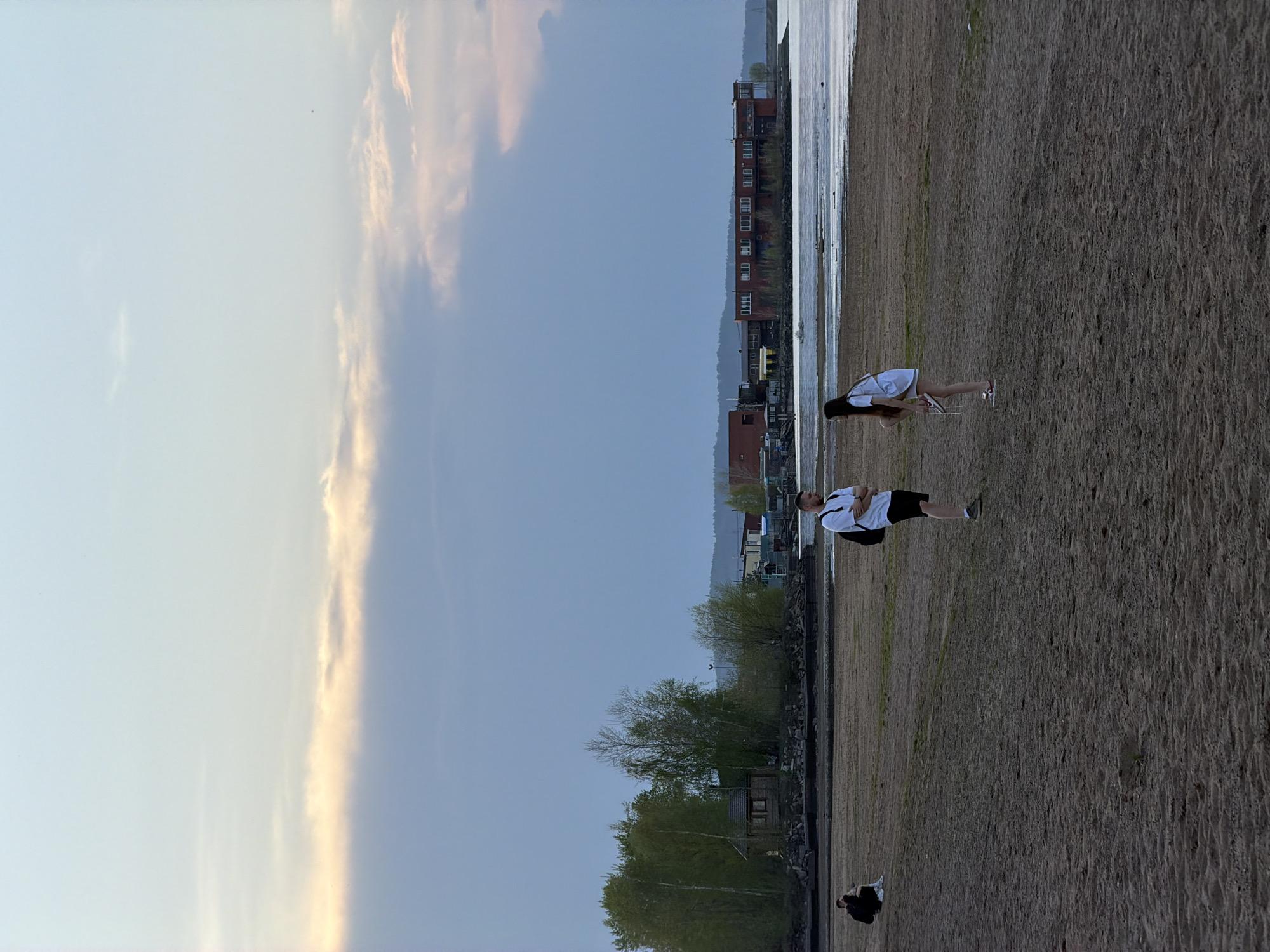 The image depicts a group of people on a sandy beach in the city of Chaikovsky, Russia. A man in a white shirt and black shorts stands on the beach. Nearby, a woman in a white dress is bending down. In the foreground, two people are sitting. The background includes buildings, trees, and a body of water. The sky is visible, with clouds tinged with a warm color.