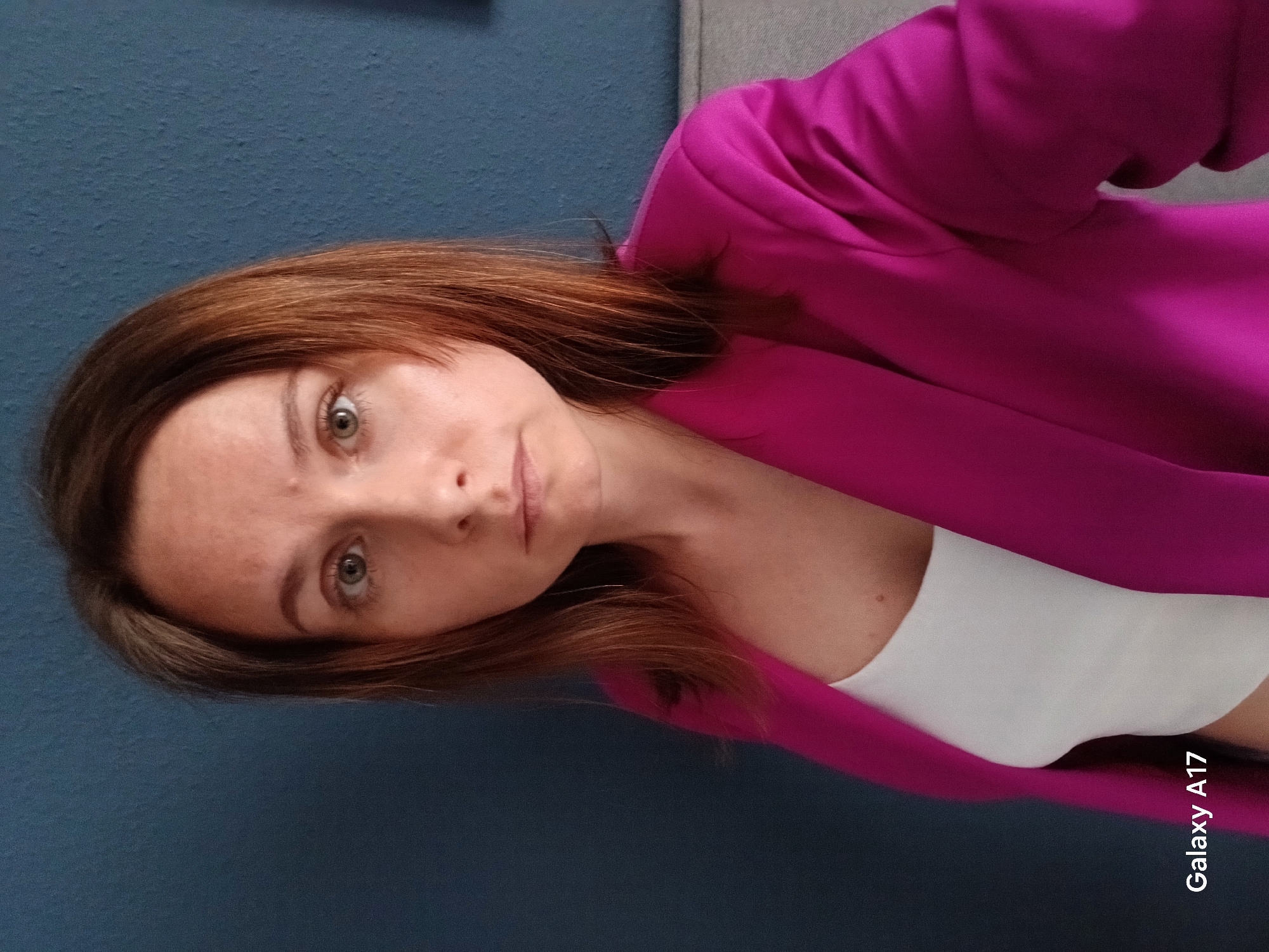 A woman with shoulder-length brown hair and green eyes is facing the camera. She is wearing a bright pink blazer over a white top. The background is a dark blue wall. The image appears to have been taken with a phone, as indicated by the watermark "Galaxy A17." There is no indication of a location in Yekaterinburg, Russia.