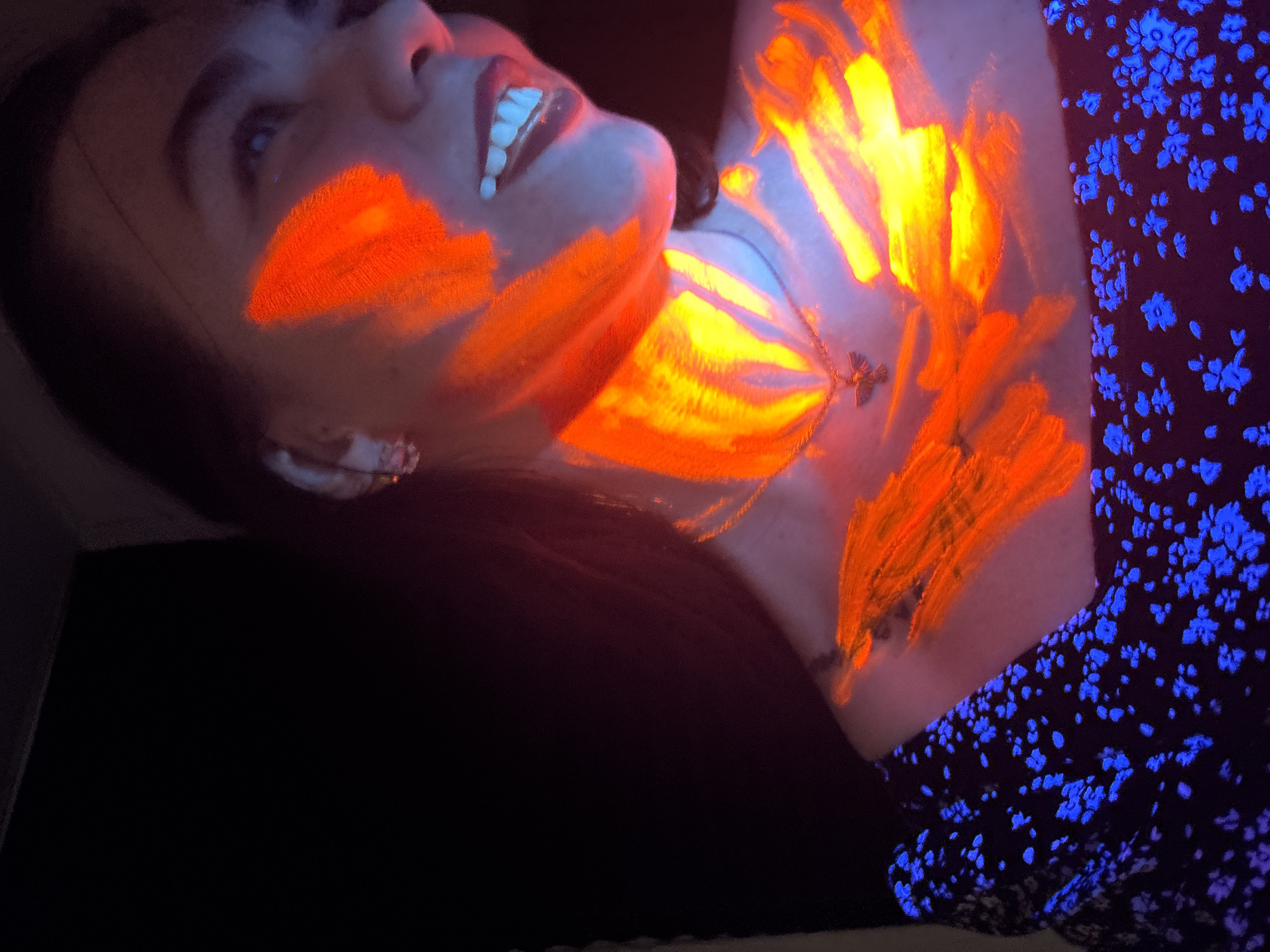 The image captures a person, likely female, in a dark indoor setting illuminated by blacklight, possibly in Perm, Russia. The individual's face and upper body are adorned with vibrant, fluorescent orange and yellow paint. On one cheek, the paint forms a distinct shape, perhaps resembling a butterfly or wings. On the neck and shoulder area, the paint is applied in abstract, brushstroke patterns that glow intensely, creating a fiery appearance.
The person's mouth is slightly open, revealing teeth, and her eyes are looking upwards, conveying a sense of expressiveness or enjoyment. She is wearing a dark-colored garment, possibly a top or dress, which features numerous small, blue floral patterns that fluoresce brightly under the UV light. A delicate necklace with a small, light-colored pendant is visible on her chest, beneath the glowing paint. The strong blacklight effect makes the painted areas and the floral patterns stand out dramatically against the dark background and the slightly purplish hue of her skin. There is no visible text in the image.
