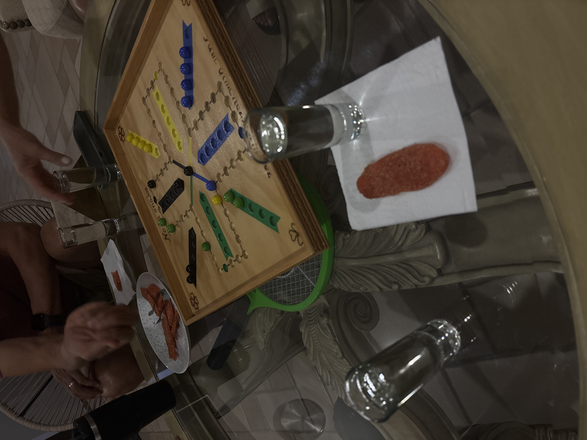 The scene captures a moment of leisure and social interaction indoors. A person seated near a light-colored, textured chair is seen with their hands near a white plate filled with reddish-brown snacks. A wooden board game with colorful pegs sits on the table along with two glass bottles filled with liquid and a piece of reddish-brown food on a white napkin. The table is round and appears to be made of glass, allowing a view of the ornate carvings on the table's base. A green tennis racket is visible beneath the glass surface. Based on the available information, I cannot determine the specific location within Chilpancingo, Mexico.