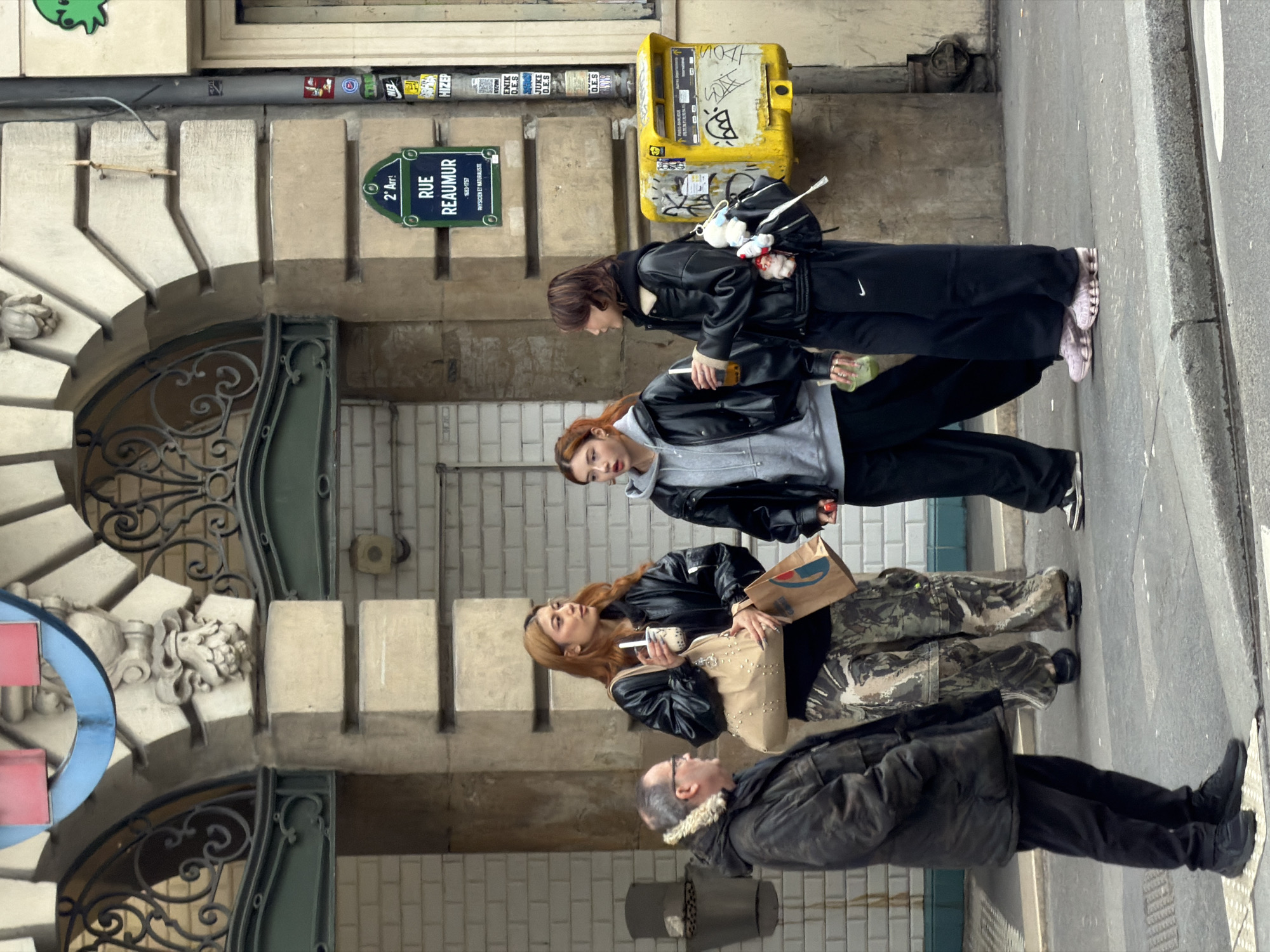 The image displays an urban street scene on Rue Réaumur, located in the 2nd Arrondissement of Paris, France, as indicated by a prominent street sign.
Four individuals are positioned on a paved sidewalk in front of a stone building facade. The building features an arched entrance with ornate dark green ironwork and visible white subway tiles within, suggesting a metro station entry. To the right, a yellow "La Poste" mailbox, heavily defaced with black graffiti, is affixed to the building. A utility pole with multiple stickers stands beside the mailbox.
On the left, an older male, dressed in a dark, possibly olive green, winter coat with a fur-lined collar, dark pants, and black athletic shoes, stands facing right, looking towards the other three individuals.
Three younger females are grouped together on the right side of the frame, holding clear plastic cups with straws containing various beverages.
The leftmost female in the group has long reddish-brown hair and wears a black bomber jacket over a dark top, paired with camouflage cargo pants. She carries a light beige studded shoulder bag and a brown paper bag. Her cup contains a milky white beverage.
The middle female has shoulder-length reddish-brown hair and wears a grey hooded sweatshirt beneath a black jacket, with black wide-leg pants and black and white athletic shoes. She holds a small red object in her right hand and a cup with a dark-colored beverage.
The rightmost female has short dark hair and is wearing a black jacket, black wide-leg sweatpants with a white Nike logo, and distinctive light pink athletic shoes with columnar soles. She carries a black shoulder bag adorned with multiple white and red plush charms and holds a cup with a light green beverage.
The three women are standing in a casual grouping, possibly conversing.