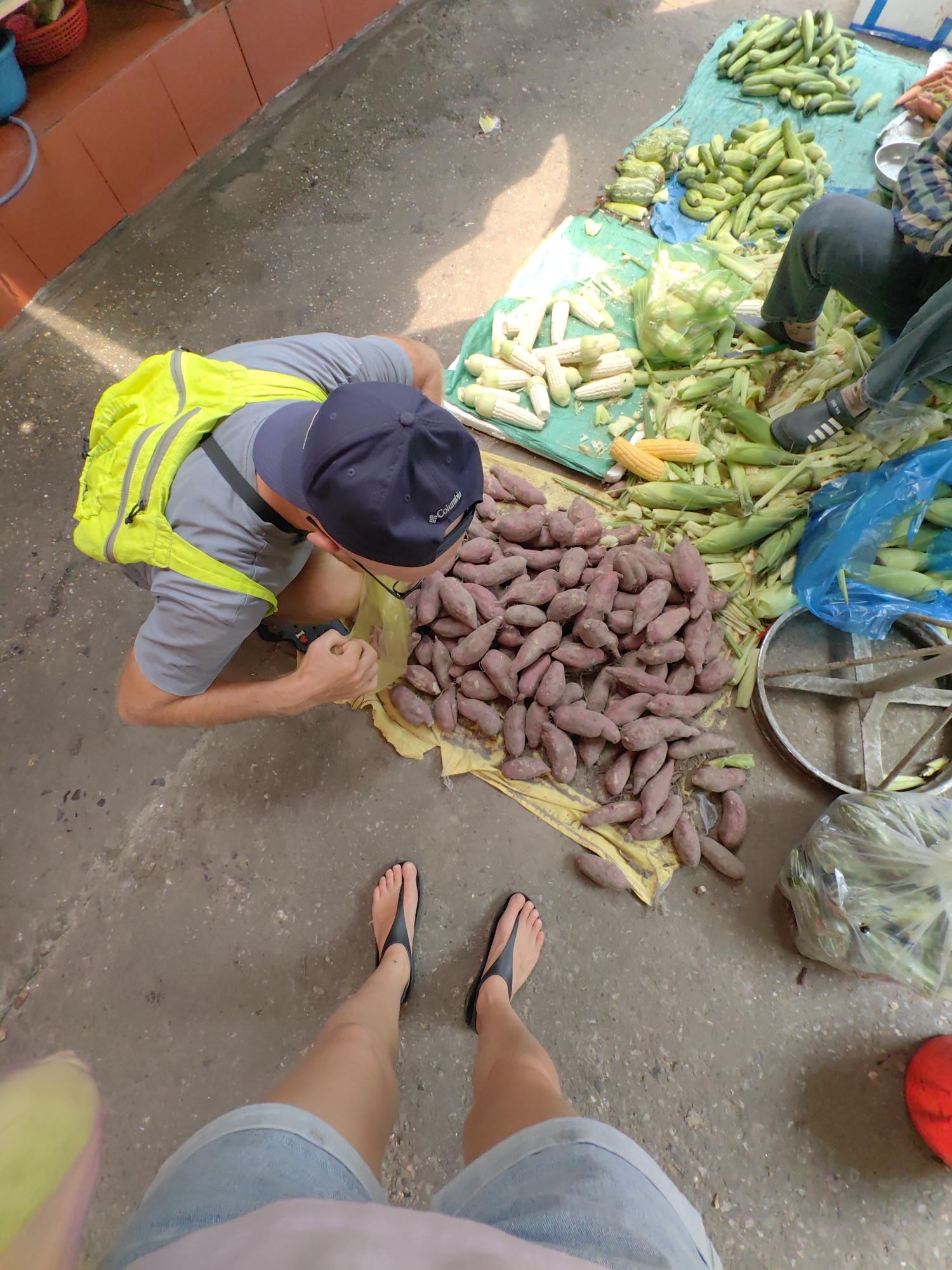 The image shows a street-level view, presumably in Ha Long Ward, Vietnam. A man wearing a gray shirt, navy blue Columbia baseball cap, and a bright yellow backpack is squatting and examining a pile of purple sweet potatoes laid out on a yellow cloth. Adjacent to the sweet potatoes are piles of corn on the cob, both with and without husks, and green cucumbers. A person's legs and feet are visible in the foreground, wearing blue denim shorts and black flip-flops. The ground appears to be concrete and slightly dirty. There is a vendor sitting with their legs visible among the produce. A bag of corn and a red object are near the feet of the person in the foreground.