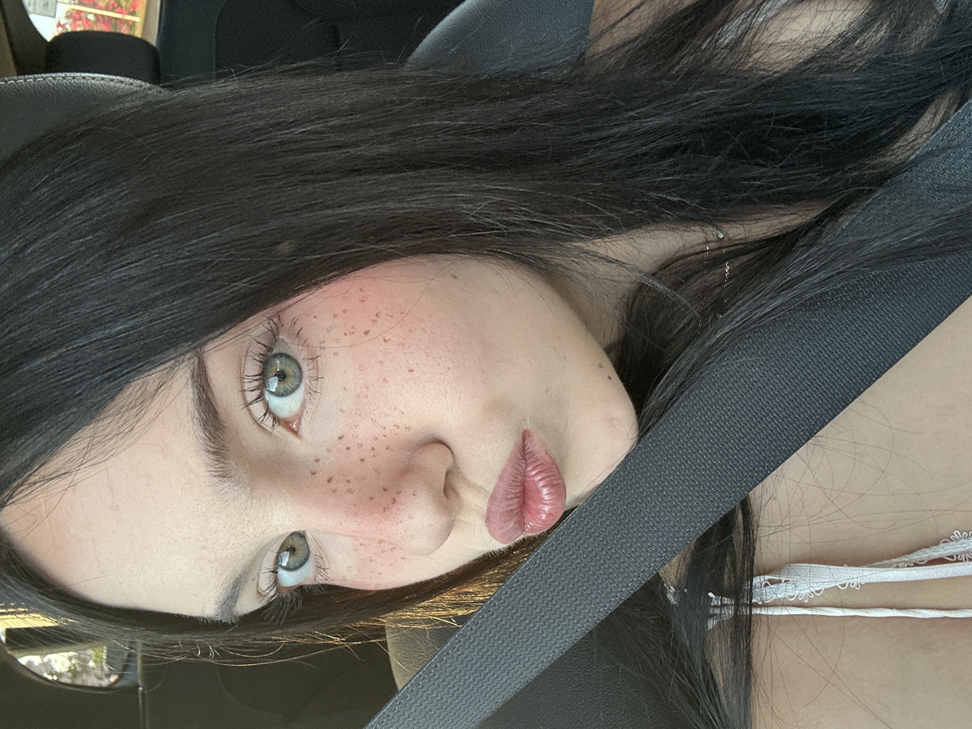 A medium close-up shot shows a young woman with dark hair and light skin, wearing a white tank top, sitting in a car. Her eyes are green, and she has freckles across her nose and cheeks. She is puckering her lips and looking towards the camera. A dark grey seatbelt crosses her chest diagonally from the left shoulder to the right side of the frame. The background is blurred, showing a hint of greenery and red flowers, suggesting an outdoor setting, possibly in or near the city of Grottammare, Italy, given the context of the prompt, although the image itself does not explicitly confirm this location. The lighting suggests it is daytime.