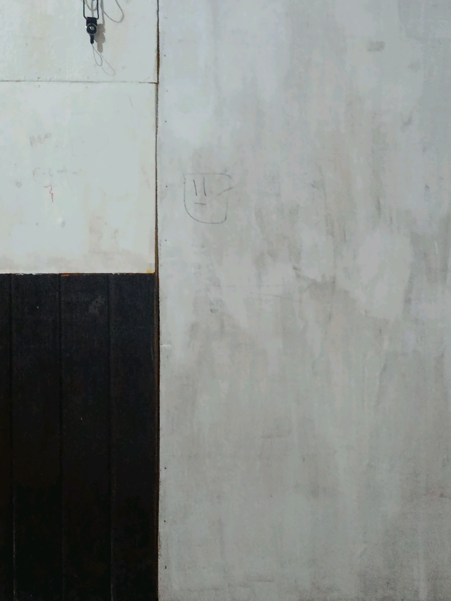The image depicts a section of an interior wall, possibly in a room or hallway. The wall is divided vertically into two distinct textures. The left side is composed of white panels, appearing to be made of a smooth material like drywall or laminate, with faint scribbles and markings on its surface. A dark wooden paneling runs along the bottom of this section.
The right side of the wall is a textured, off-white surface, possibly plaster or concrete, with visible stains and imperfections, suggesting age or lack of recent maintenance. A child-like drawing of a smiley face is visible on this textured section. At the top of the image, a small black object with wires hanging from it is attached to the wall, its purpose unclear.
This image is located in Loa Janan Ulu, Indonesia.