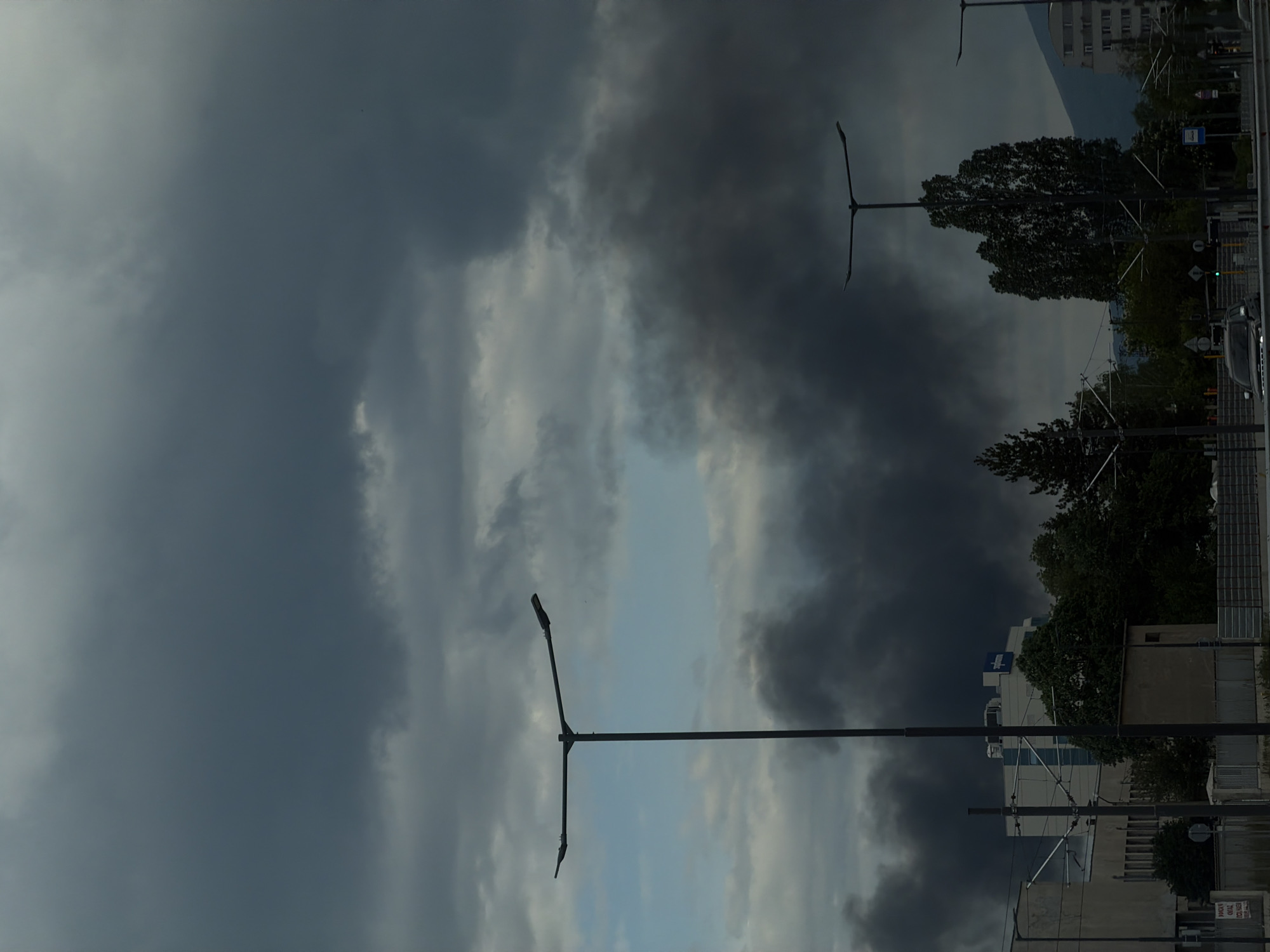 The image depicts a street scene in Sofia, Bulgaria under a stormy sky. Dark, heavy clouds dominate the frame, with a small patch of light blue sky visible. Streetlights with a "Y" shaped design are prominent in the foreground. The background features buildings, trees, and a mountain range partially obscured by the clouds. There are power lines present. The lighting suggests overcast conditions.