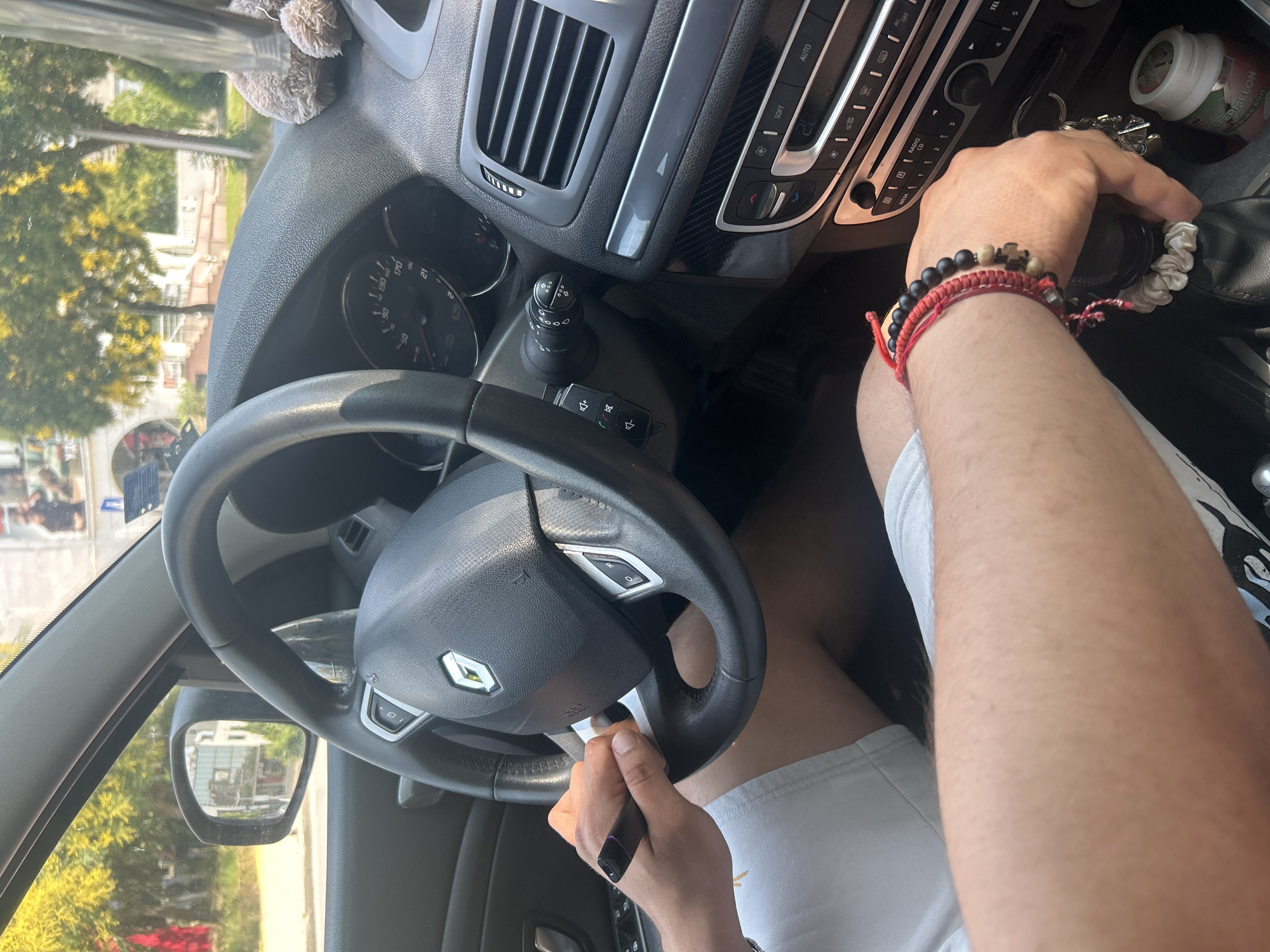 The image shows the interior of a car, likely a Renault, in Plovdiv, Bulgaria. A person's hands are visible, one holding a black vape pen and the other resting on the gear stick. The person is wearing white shorts and has bracelets on their wrist. The car's dashboard is visible, including the steering wheel, air vents, and radio. Outside the car, trees and buildings can be seen, suggesting an urban environment. The sunlight indicates it is daytime.