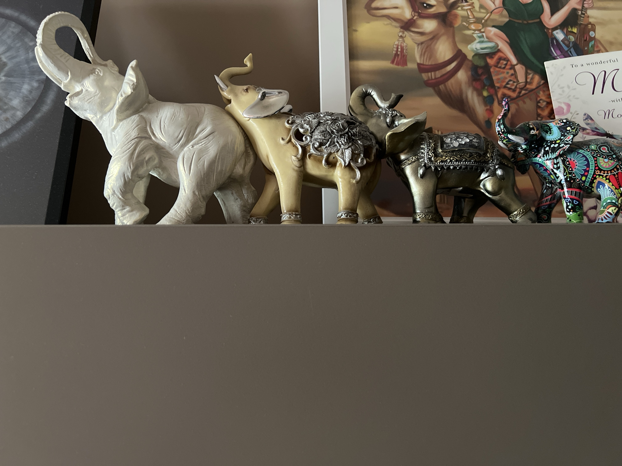 The image shows a collection of five elephant figurines arranged on a shelf. From left to right, the elephants are white, pale yellow, brassy gold, darker gold, and multi-colored (black, blue, red, green). A portion of a framed print is visible behind the elephants. A card or note with text is also visible behind the elephants. The shelf is a neutral brown color, as is the wall behind it. No people are visible in the image. There is no indication of the location of the image (Burgas, Bulgaria).