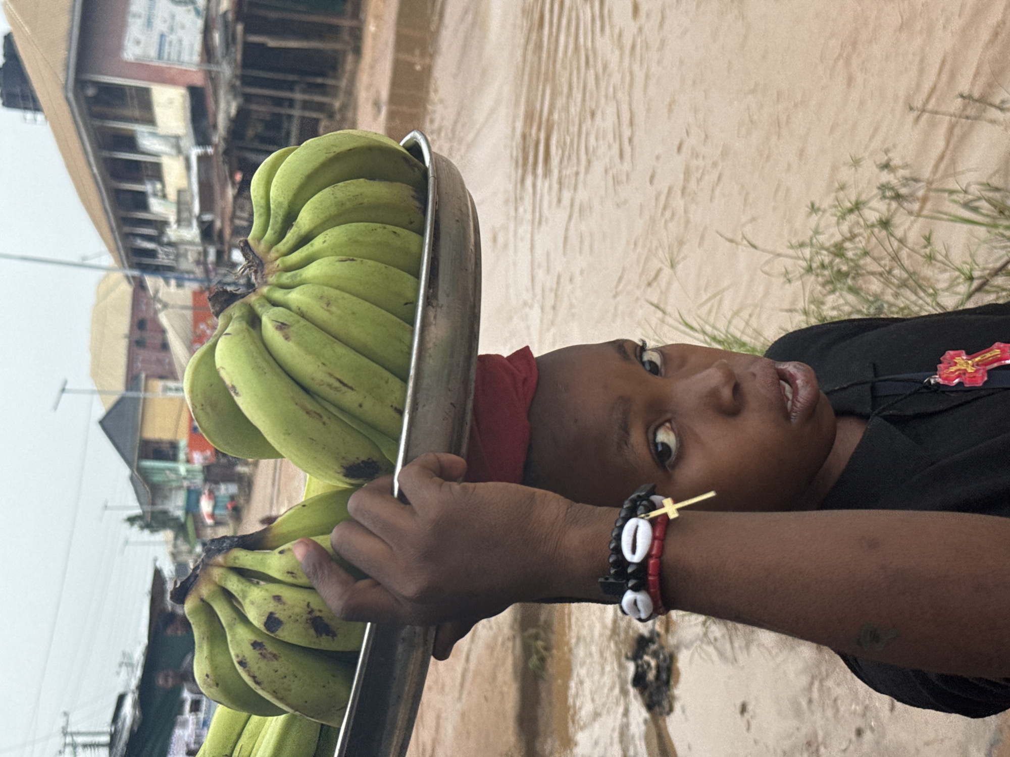 In Ihiagwa, Nigeria, a young person carries a metal tray laden with green bananas balanced on their head. The person is looking directly at the camera with a slightly open mouth. They are wearing a black collared shirt and a red cloth is visible around their head, under the tray. A bracelet with black, white, and red beads and a golden cross charm is worn on their visible arm. The background shows a street scene with buildings and a body of muddy water.