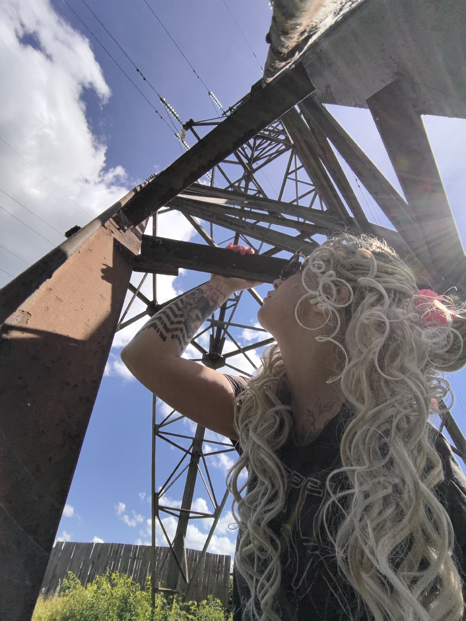 An overhead low-angle shot shows a young woman with voluminous, curly blonde hair and dreadlocks, wearing sunglasses and a dark t-shirt with a graphic print. She is standing outdoors, looking up at a towering metal power line pylon under a bright blue sky with scattered white clouds. Her left arm is extended, touching the pylon's rusty metal structure. A large tattoo is visible on her forearm. The pylon is a complex lattice of steel beams. In the background, a weathered wooden fence runs horizontally, bordered by green vegetation. The scene is bathed in sunlight, with lens flare visible from the upper right.