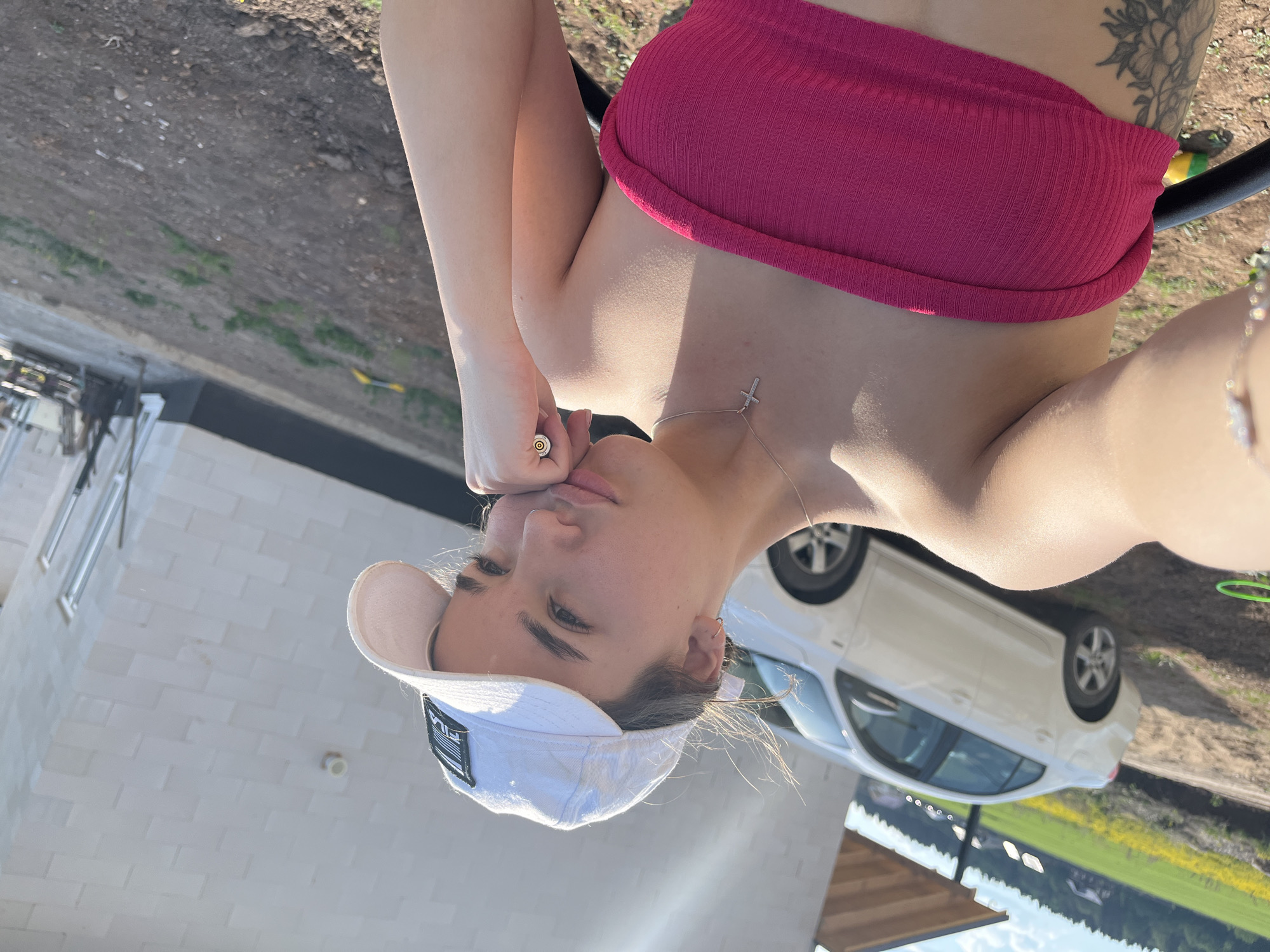 A young woman wearing a pink tube top and a white baseball cap is posing outdoors. She has a silver necklace with a cross pendant. The background includes a white car, a building under construction, and a field. The location is Лобаново, Russia.