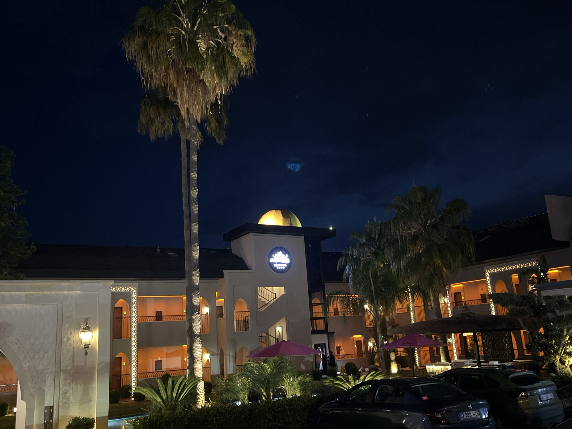 The image is taken at night in Alanya, Turkey, and shows the exterior of a light-colored building with a dome on top. A tall palm tree stands on the left. The building has multiple floors with arched windows and balconies lit with orange lights. There are various trees and plants in the foreground and several parked cars, including a dark blue one with a license plate visible. Two pink umbrellas are also visible in the middle of the image.