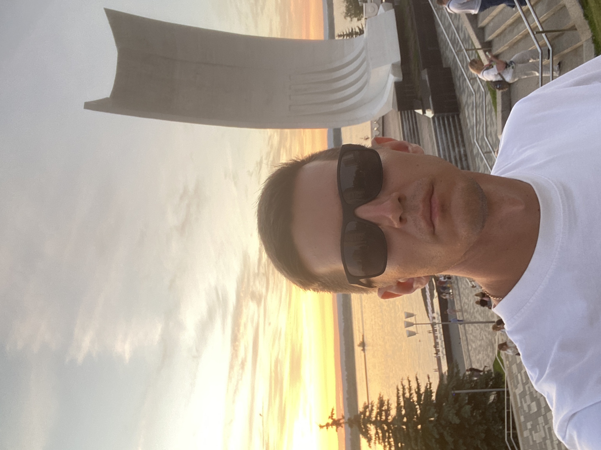 This is a selfie taken outdoors, oriented vertically. The primary subject is a man wearing sunglasses and a white t-shirt, seen from the chest up. Behind him, to his right, a large, light-colored, abstract sculptural structure with curved elements is visible. In the background, a body of water can be seen, reflecting the warm colors of a sunset. The sky is filled with soft, wispy clouds, indicating it is either dawn or dusk. The lighting suggests it is sunset, casting a warm glow over the scene.
To the right of the main subject and the sculpture, a set of stairs with metal railings is present. Several people are visible on these stairs and along a paved walkway adjacent to the water. Some of these individuals are looking towards the water or standing in groups. Trees with dark green foliage are also visible in the lower right portion of the image, bordering the paved area. The overall setting appears to be a public space near a waterfront, possibly a park or promenade. The emotion conveyed is relaxed and casual, typical of a selfie taken during leisure time. No visible text is present in the image.