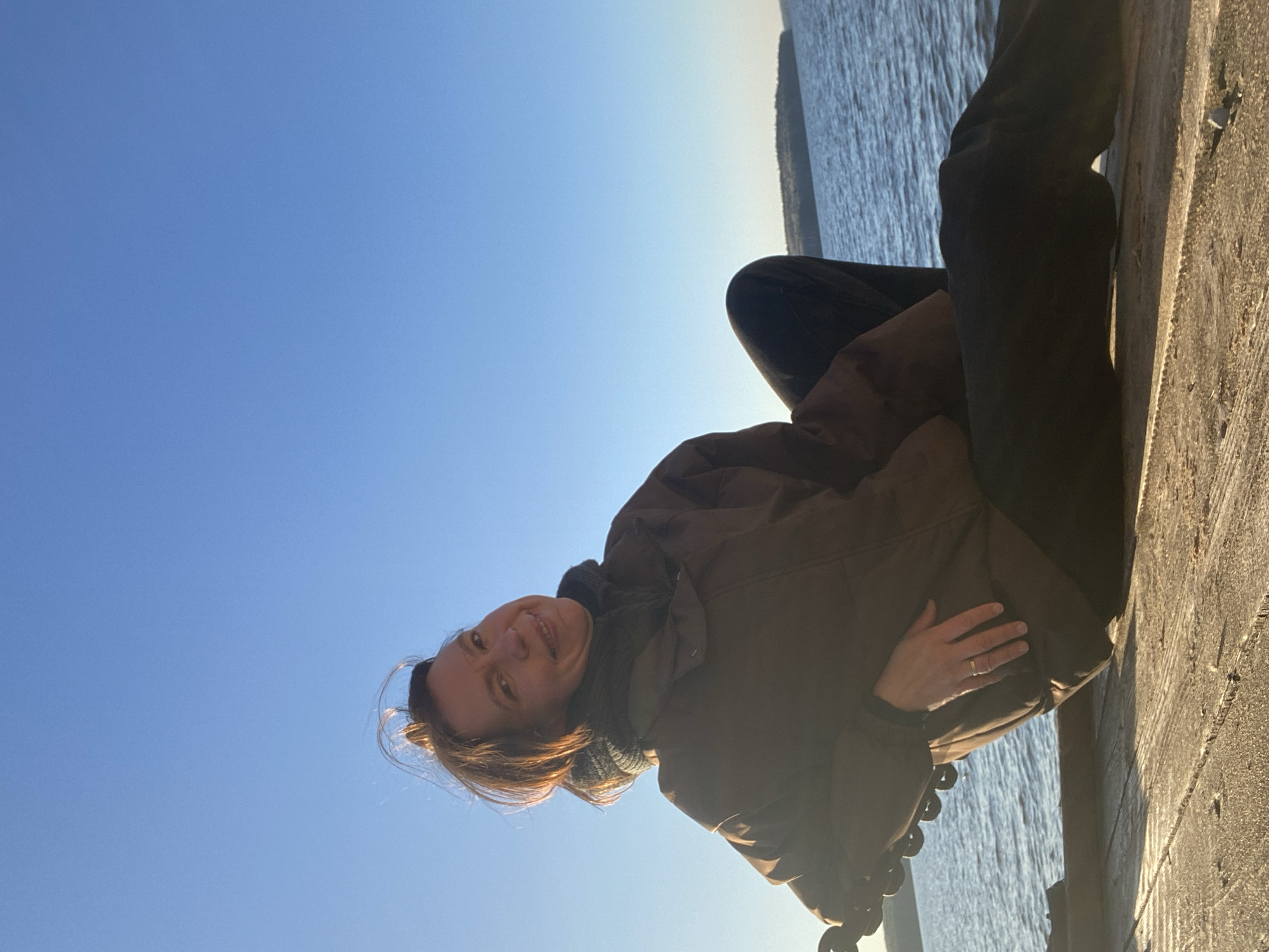 A woman with shoulder-length brown hair is sitting on a concrete ledge near water. She is wearing a brown jacket, dark pants, and has a wedding ring on her left hand. She is smiling and looking up. The sky is blue and there are signs of the city of Chaikovsky, Russia.