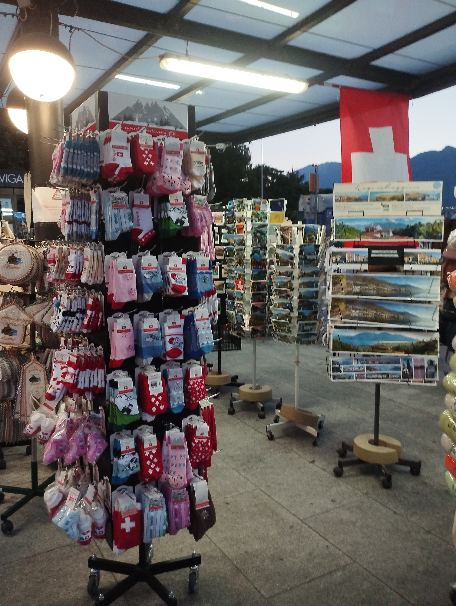 The image depicts an outdoor market stall in Locarno, Switzerland, featuring a display of Swiss-themed merchandise, primarily socks and postcards. The stall is set up under a covered walkway with overhead lighting. A large Swiss flag hangs prominently in the background.
In the foreground, several racks are filled with colorful socks. Many of the socks are white with red Swiss crosses or other patriotic designs. Other socks feature cartoon characters and patterns like hearts and flowers. The socks are neatly displayed on hangers, with some packaged in plastic.
To the right of the sock display, racks of postcards showcase scenic views of Switzerland, including landscapes with mountains, lakes, and historical buildings. The postcards appear to be arranged in horizontal rows, with each postcard displaying a different image.
The ground beneath the stall is paved with stone tiles. The overall scene suggests a tourist-oriented souvenir shop or a market catering to visitors of Locarno. The lighting indicates it is either early morning or late evening. There are no people visible in the image. The overall atmosphere is one of a commercial retail environment in an outdoor setting.
