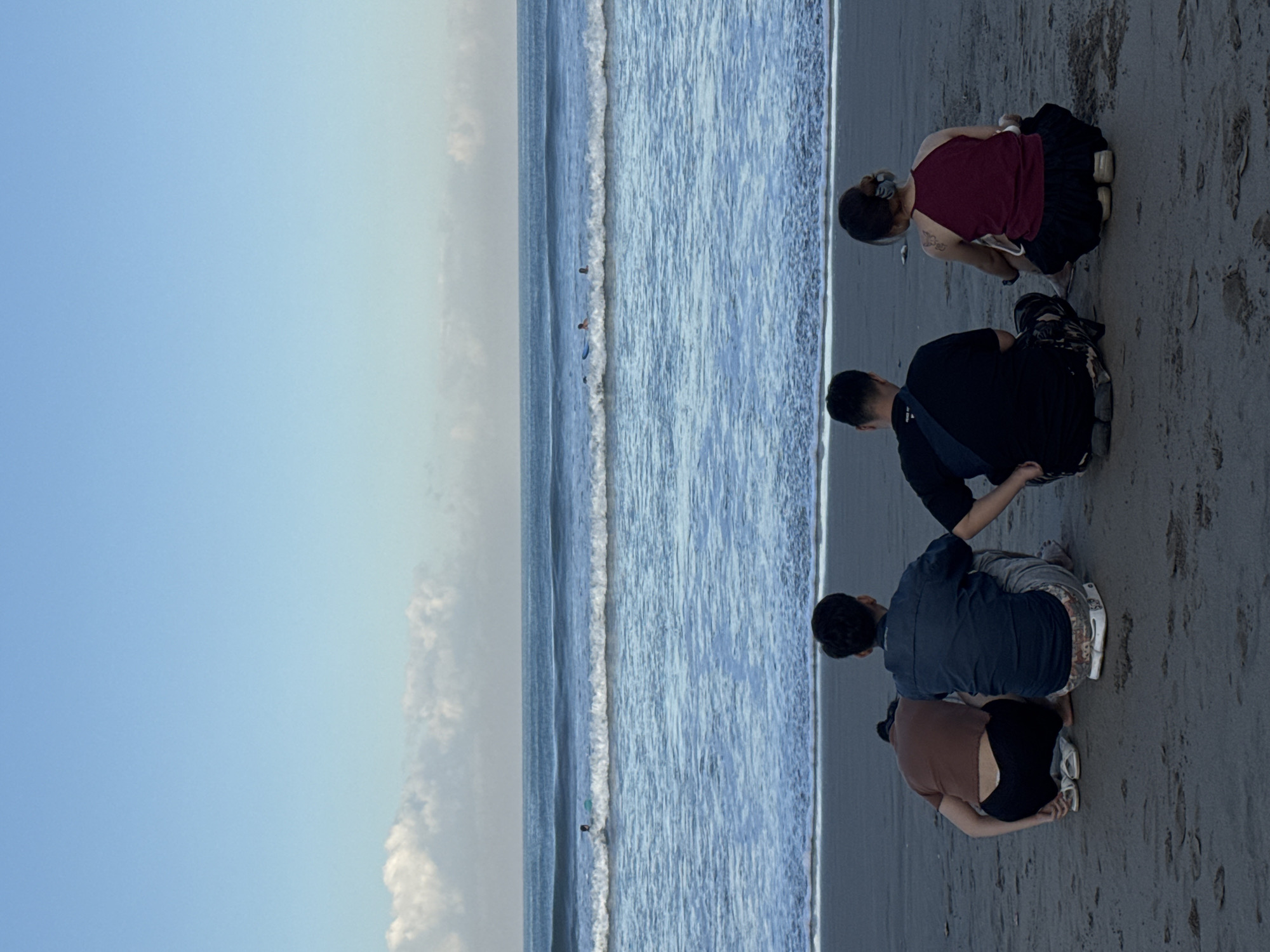 The photograph shows four individuals sitting on the beach in Kuta, Indonesia, next to the ocean. The sky is predominantly clear blue with some clouds. Several people are visible in the water further out, likely surfing. The individuals on the beach appear to be looking at something on the sand. One is wearing a maroon tank top and a dark skirt, another a black shirt and camo pants, another a blue shirt and gray pants, and the last a brown shirt and black pants. The sand is wet and gray.