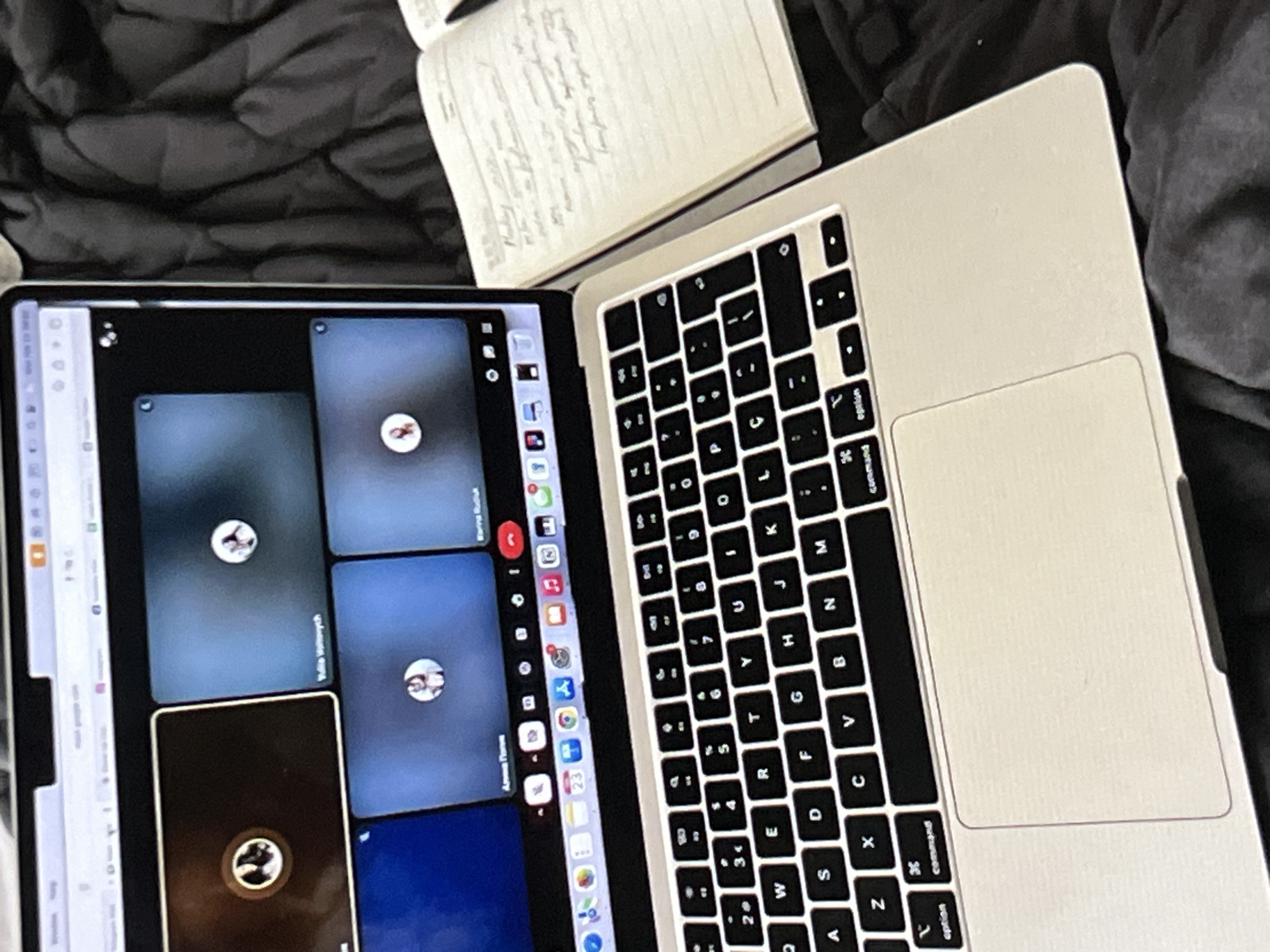 The image shows a close-up, overhead view of a laptop and a notebook on a dark, textured surface, possibly a bed or blanket. The laptop is open, displaying a video call interface with several participants' video feeds visible. The notebook, lying open next to the laptop, contains handwritten text. The scene appears to be set indoors, given the artificial lighting and the presence of electronic devices.
The focus is on the laptop screen, which shows a grid of video windows, suggesting an active online meeting or class. Each window displays a participant's profile picture or live video feed. Some of the names or labels beneath the windows are partially visible, with "Арина Попния" being the most legible. The overall mood is one of engagement with digital communication or remote work/study.
Notable details include the keyboard of the laptop, the trackpad, and the spine of the open notebook with its pages filled with writing. The lighting is somewhat dim, and the background is a dark, soft texture, contributing to a cozy or perhaps late-night atmosphere. No specific time of day or weather conditions can be inferred. The emotions of the people in the video call are not discernible due to the image quality and focus. The scene is set up for work or study, with the notebook possibly used for taking notes during the call. The visible text on the screen and in the notebook provides context for the activity.