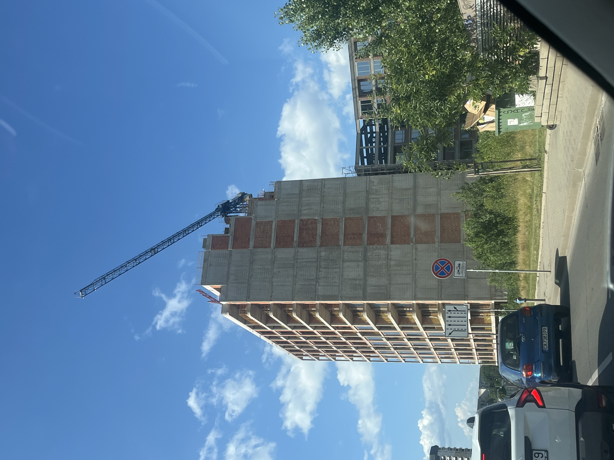 The image depicts a street scene in Sofia, Bulgaria, on a sunny day with a bright blue sky dotted with white clouds. In the foreground, a white SUV is partially visible on the left, and a blue Toyota Yaris is in front of it. Behind the cars, a tall, unfinished building under construction dominates the scene. A construction crane extends from the top of this building. To the right of the unfinished building, another building, also appearing to be under construction with exposed concrete framework, is visible. Further to the right, dense green trees and a large green dumpster filled with cardboard boxes are present. Traffic signs are also visible, including a sign indicating "No stopping or parking" and a traffic direction sign. The overall impression is one of urban development and ongoing construction.