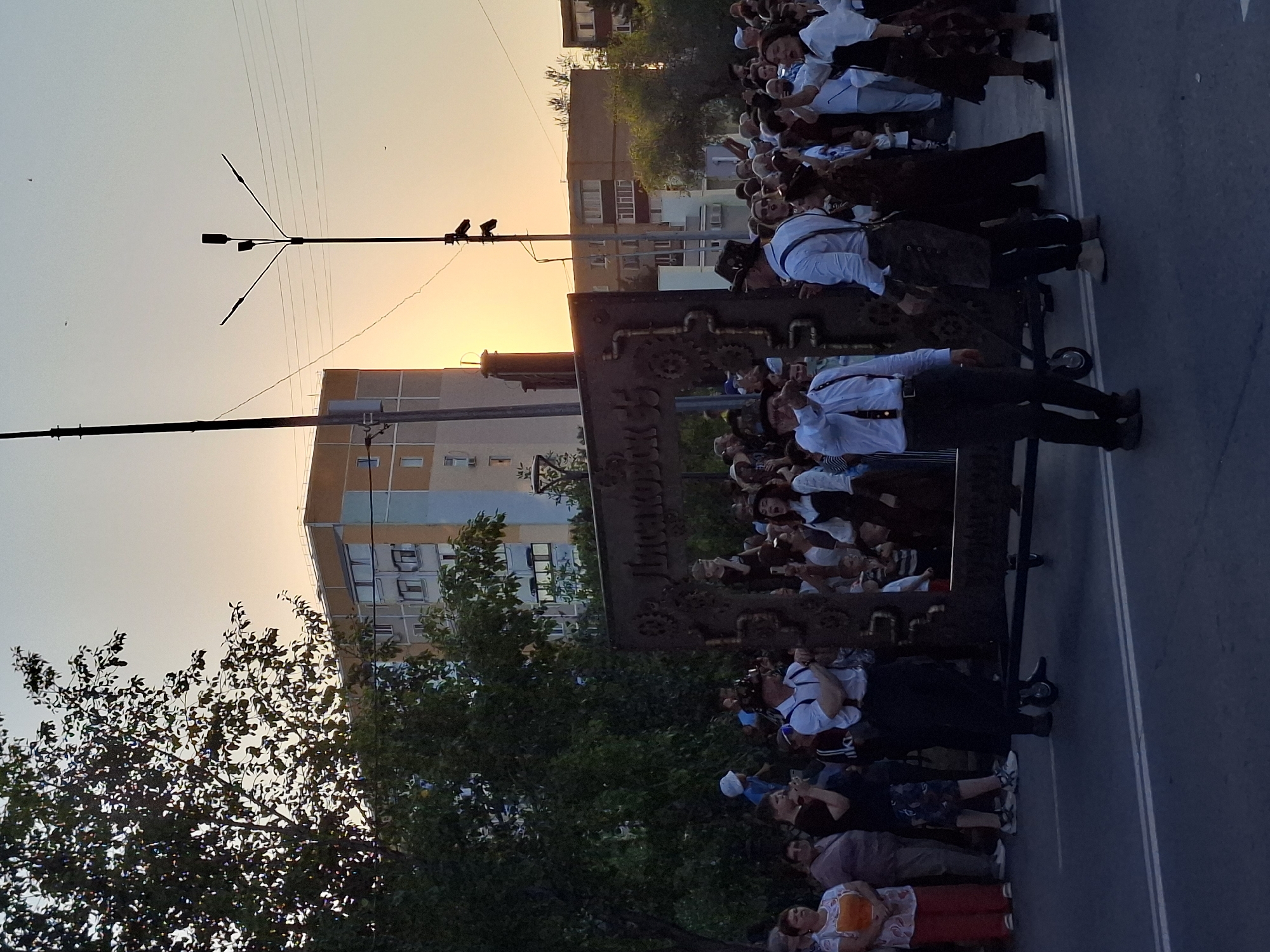 A large group of people, many dressed in steampunk-inspired attire, gather on a street as the sun sets, casting a warm glow. In the center of the frame, a large, ornate metal structure, possibly a decorative archway or prop, is being moved by people. The structure features intricate gears and pipes, resembling a steampunk aesthetic. Buildings, including a multi-story apartment complex, line the street in the background, some lit by the setting sun. A streetlight with multiple bulbs stands tall against the sky. The atmosphere appears festive, with some individuals interacting with the camera and others observing the scene. The ground is asphalt with visible lane markings.