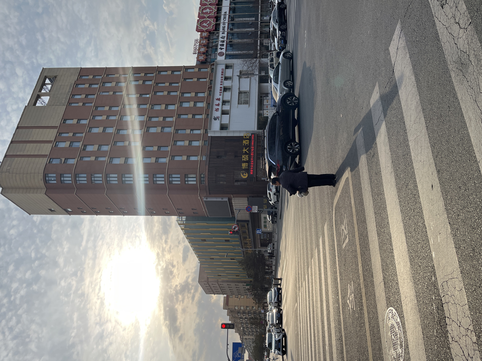 A street scene in Changchun City, China, features a tall, light brown building to the left, reaching into a partly cloudy sky where the sun is shining brightly. A row of parked cars lines the side of the road near the building, and a person is crouched down on a white-striped crosswalk. Other buildings are visible further down the street, along with a traffic light that is red. Signs written in Chinese characters are present on several of the buildings.