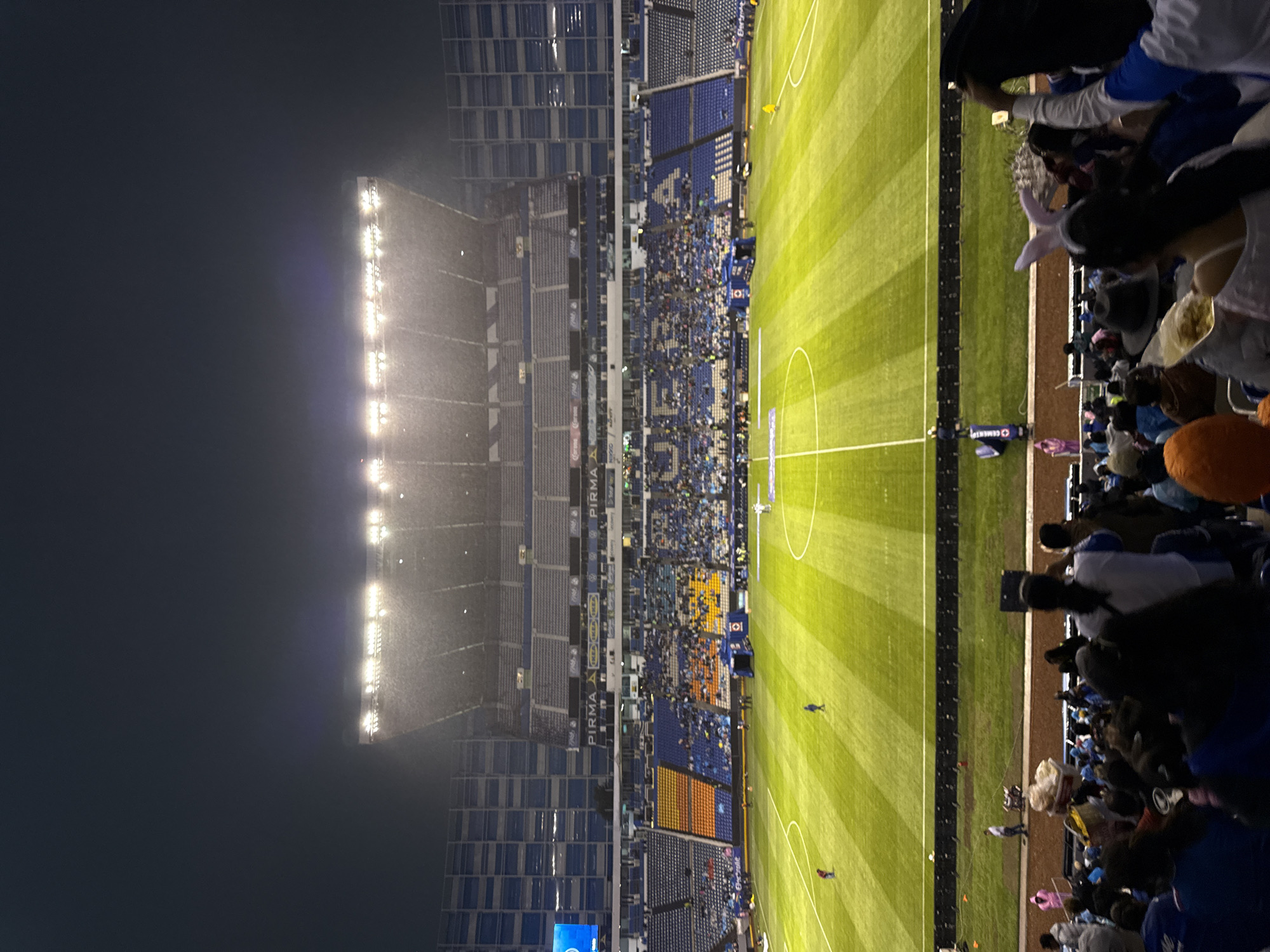 The image shows a soccer game being played in Puebla, Mexico. The stadium is crowded with fans, and the field is well-lit. A heavy mist or light rain is falling near the stadium lights. The stands are predominantly blue with patches of orange and gray. The pitch is lined and includes a center circle. People in the stands are watching the game.