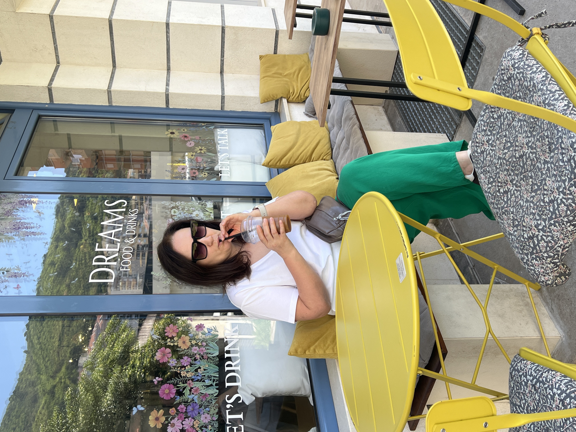 A woman with long, dark hair and sunglasses sits at a yellow metal table outside the "Dreams Food & Drinks" cafe in Budva, Montenegro. She is wearing a white t-shirt and is seen drinking from a clear plastic cup with a straw. She is seated on a cushion on a bench with yellow pillows behind her. The cafe window has floral decorations and text that includes "Dreams Food & Drinks" and "Let's Talk." The scene takes place during daylight.