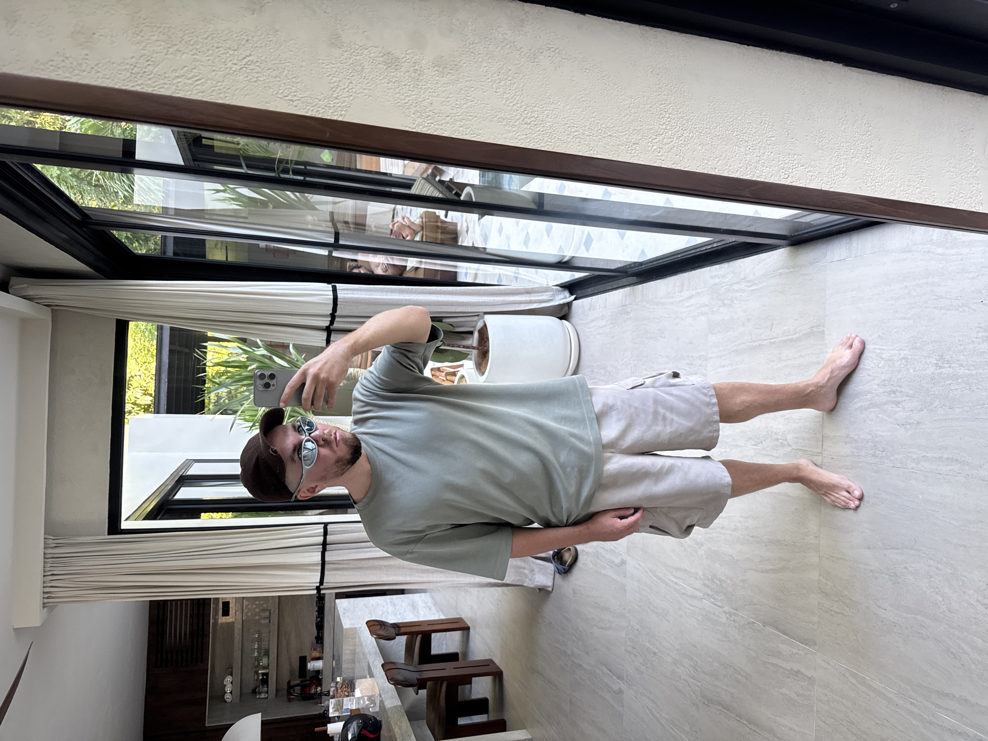 A young man is captured in a full-body selfie, standing barefoot inside a modern building with large glass windows overlooking a tropical outdoor setting. The image is taken as if the man is standing upright, with the floor to his right. He wears a light olive green short-sleeved t-shirt, light beige cargo shorts, a brown baseball cap, and light-framed sunglasses. He holds a grey smartphone up, seemingly taking the picture, with part of his arm visible at the top of the frame.
The man stands on a light beige or grey marble-patterned tiled floor. To his left, partially drawn white sheer curtains cover the bottom portion of tall, black-framed glass windows or sliding doors. Through these windows, lush green tropical foliage and bright daylight are visible, suggesting a sunny day. A large white cylindrical planter with a plant is positioned near the windows.
In the background, behind the man and through the reflections on the glass, a modern interior space is visible. This includes two dark wooden bar stools with curved seats, and a counter area that appears to be a bar, stocked with various bottles and glassware. A pair of dark sandals or flip-flops rests on the floor near the man's feet. The ceiling is a textured white. The overall impression is a contemporary, light-filled space, likely in a resort or home, consistent with the Kuta Selatan, Indonesia location context. There is no visible text in the image.