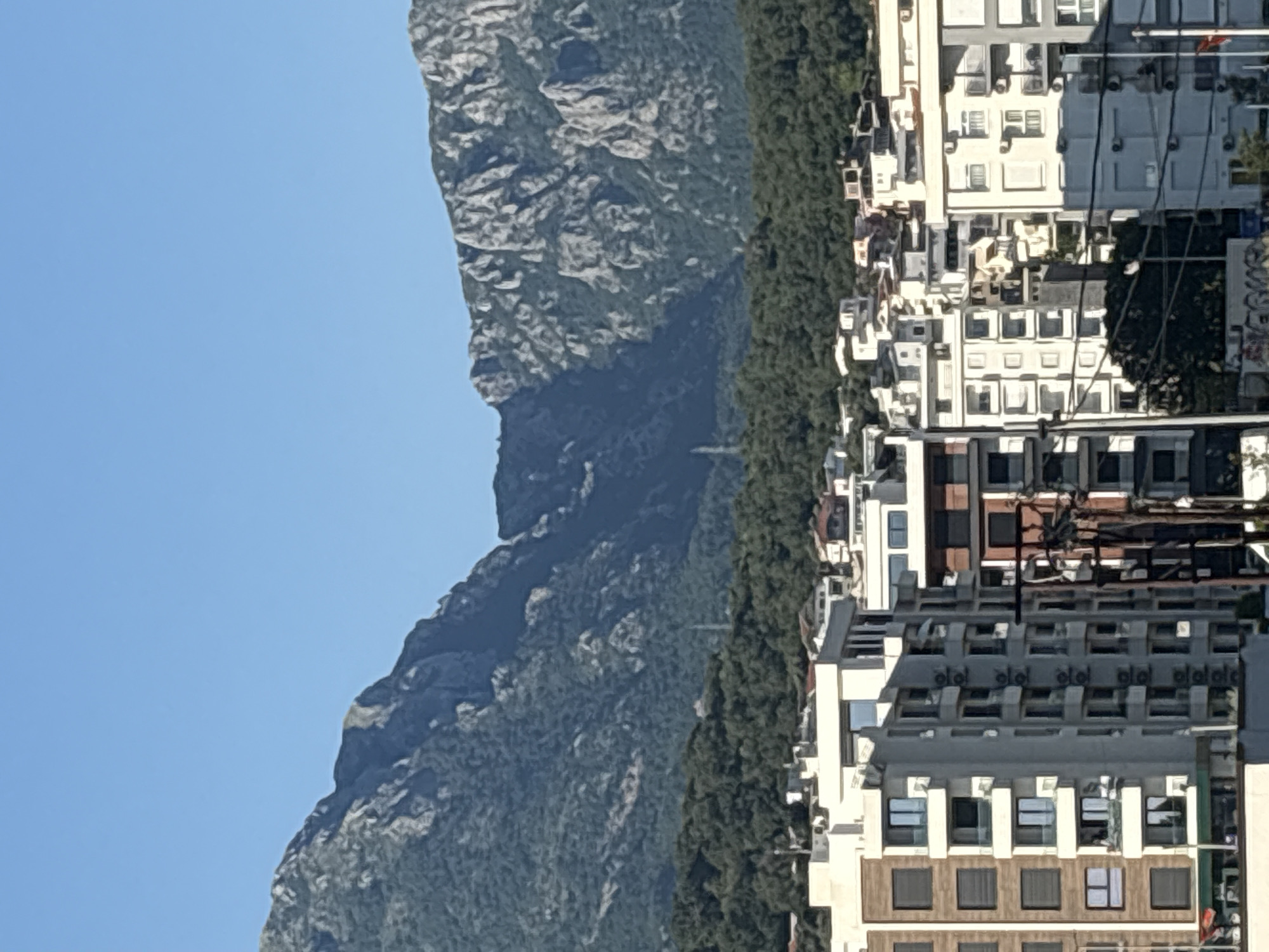 The image captures a detailed view of the city of Bar, Montenegro, with the prominent Rumija Mountain range forming the backdrop under a clear, bright blue sky. The mountains exhibit rugged, grey rocky peaks and exposed crags, interspersed with extensive patches of dense, dark green vegetation covering their lower slopes and extending into the foothills. A slender, light-colored antenna or utility tower is visible on the densely wooded mountain slope in the mid-ground, slightly to the left of the center.
The foreground is occupied by a dense cluster of modern multi-story buildings, predominantly residential, constructed with light-colored facades in shades of white, cream, and grey. One building on the far left features a distinctive brown and white striped facade. Many buildings display numerous balconies and large windows. A high density of external air conditioning units is visible on the balconies and exterior walls of several buildings, particularly a light-grey structure in the center-right foreground. Utility poles and dark power lines traverse the immediate foreground, partially obscuring the lower sections of some buildings. No individuals are visible. The scene is illuminated by strong sunlight, indicating clear daytime conditions. A small red flag or banner is barely discernible on a structure in the lower right corner, and some faint graffiti is present on a low wall in the bottom center.