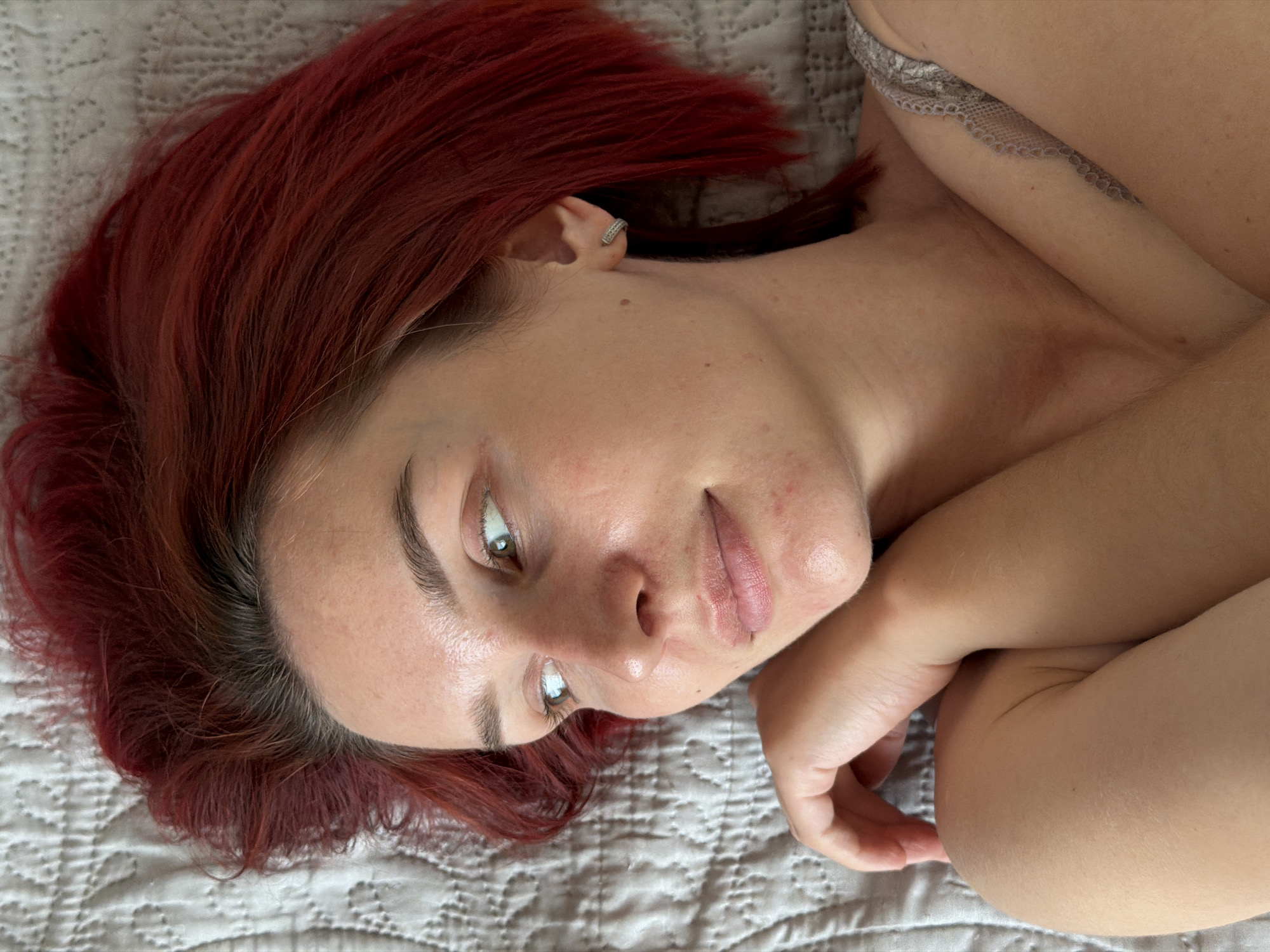 A young woman with red hair is lying on a light-gray quilted bedspread. She is fair-skinned, with light-brown eyes and visible freckles and blemishes on her face. She wears a silver-colored earring in her left ear. Part of a patterned garment, possibly lingerie, is visible at the top right corner of the image. Her hands are folded near her chest. The location, Chaikovsky, Russia, cannot be confirmed or denied with the information provided in the image.