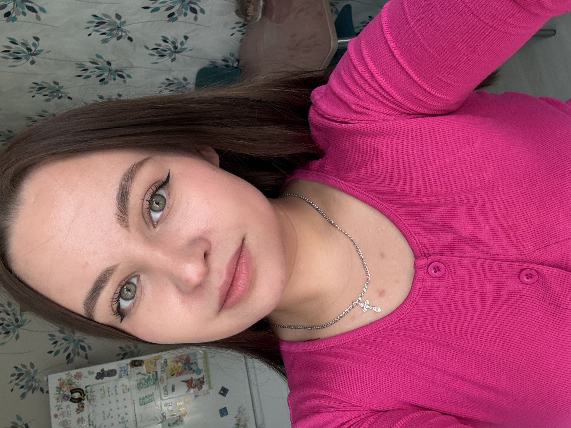 A young woman poses for a selfie indoors, likely in a private residence. She has brown hair, green eyes, and is wearing winged eyeliner. She is also wearing a bright pink, ribbed-texture top with buttons. A silver necklace with a small cross pendant is visible around her neck.
The background includes a patterned bedspread and a refrigerator covered with stickers. A chair is visible further back. The location is specified as being in Марковский, Russia.