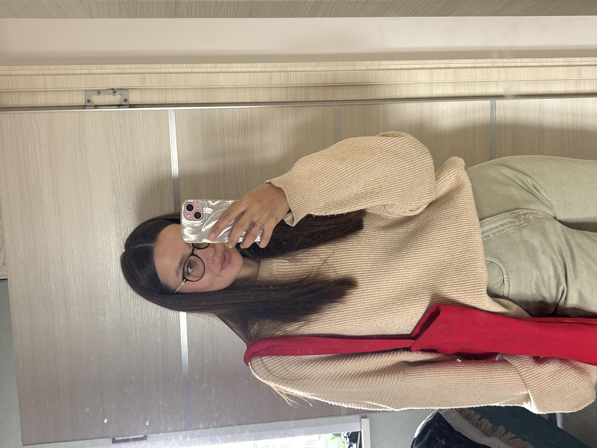 A woman is taking a mirror selfie in a room. She has long, dark hair, wears round glasses, and is dressed in a beige knitted sweater and light green pants. A bright red tote bag is slung over her left shoulder. The background consists of a mirrored closet door and a section of a wall. The location is identified as Krasnoyarsk, Russia.