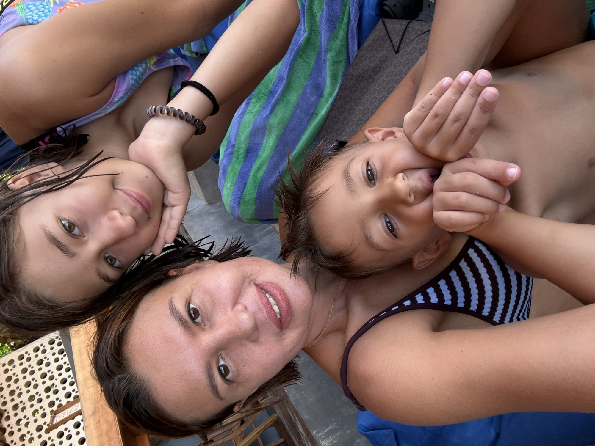 A selfie photo of a woman and two children, possibly on a beach in Kuta Selatan, Indonesia, shows them laying down and smiling. The woman is at the bottom of the frame, with short brown hair and a necklace. She is wearing a black and blue striped bathing suit. To her left is a girl with long brown hair and a bracelet, wearing a blue bathing suit with pineapples. To the right of the woman is a shirtless boy with brown hair, whose hands are raised near his face. A blue and green striped towel is visible in the background. A wooden structure is on the left edge of the frame.
