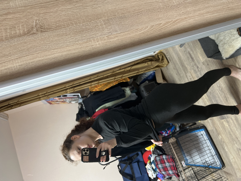 The image displays a mirror selfie of a person, likely a young woman, captured in what appears to be an indoor room. The photo is rotated approximately 90 degrees to the right, so the person is horizontally oriented across the frame, facing the left side of the image.
The person has curly, reddish-brown hair and is looking towards the camera with a neutral expression. She is holding a black smartphone with a distinctive case that has white or light-colored rectangular patterns on the back. Her fingers, painted with pink nail polish, are visible gripping the phone. She is wearing a long-sleeved black top and form-fitting black leggings, which appear to have a slight shimmer or sparkle. One hand is holding the phone, while the other hand is slightly tugging at the bottom edge of her top, revealing a sliver of her midriff.
The background, which is the reflection in the mirror, shows a cluttered room. The floor is made of light-colored wood planks. In the upper right corner of the reflected scene (which is the bottom right of the image), there's a dog crate or pet carrier made of metal bars with a white tray inside. To the right of the crate, a fluffy, off-white cushion or blanket is visible on what might be a pet bed or a piece of furniture.
Various items of clothing and bags are scattered on the floor and possibly draped over furniture. A dark blue duffel bag is prominent in the lower left of the reflected scene. Near it, there's a red object, possibly a hat, and a plaid shirt with red, black, and white squares. In the mid-ground, a rolled-up, light brown or golden-colored curtain or blind hangs, and beneath it, some framed pictures or posters are partially visible on a surface. The overall impression of the room is that it is lived-in and somewhat untidy. No discernible text is visible in the image.