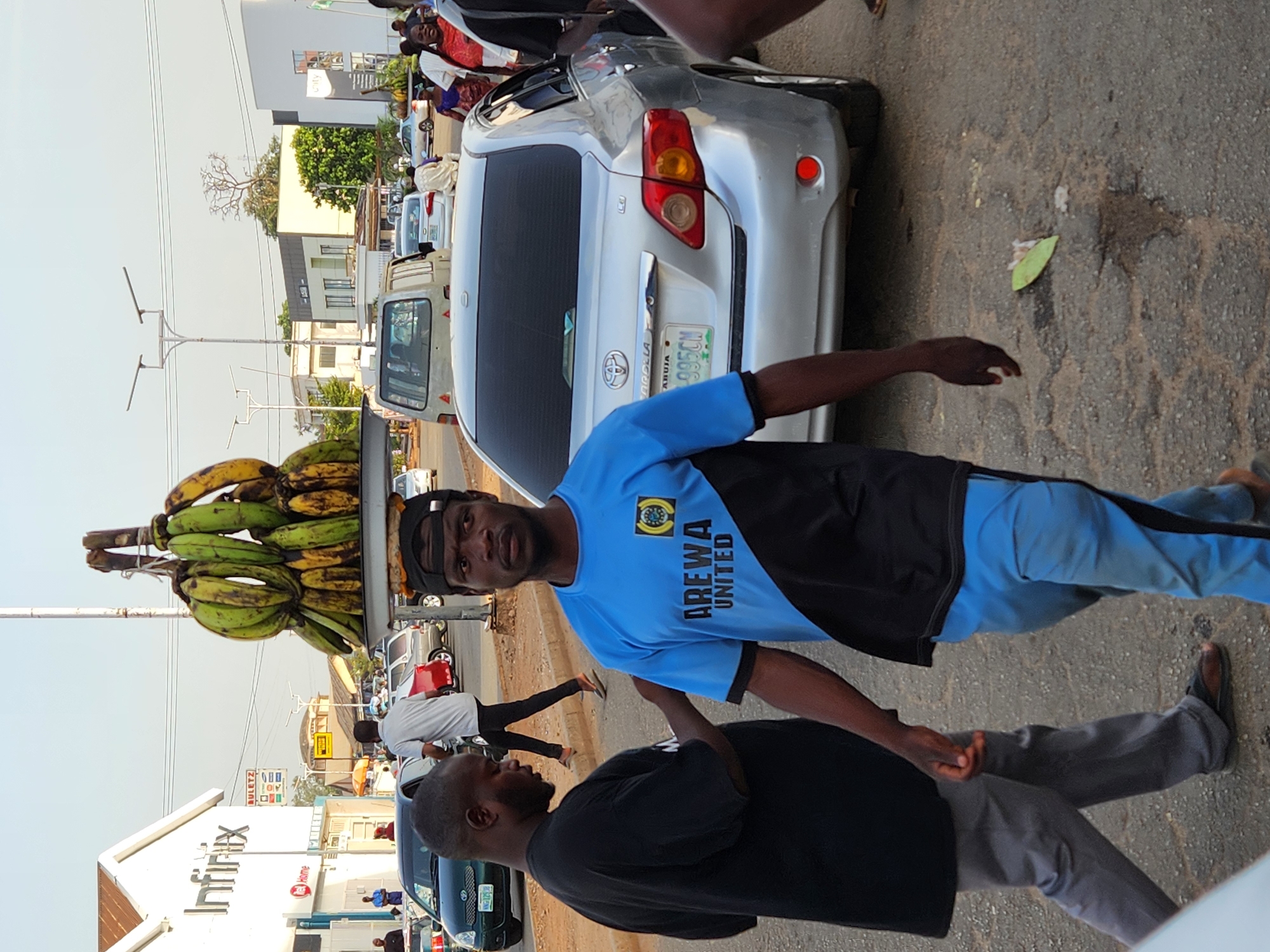 A man in a blue and black "Arewa United" t-shirt carries a tray of unripe bananas on his head on a street in Jos, Nigeria. Another man in a black t-shirt stands beside him. They are on a paved street alongside a silver Toyota Corolla with Abuja plates and other vehicles. An Infinix store is visible in the background. There are other people and shops in the distance.