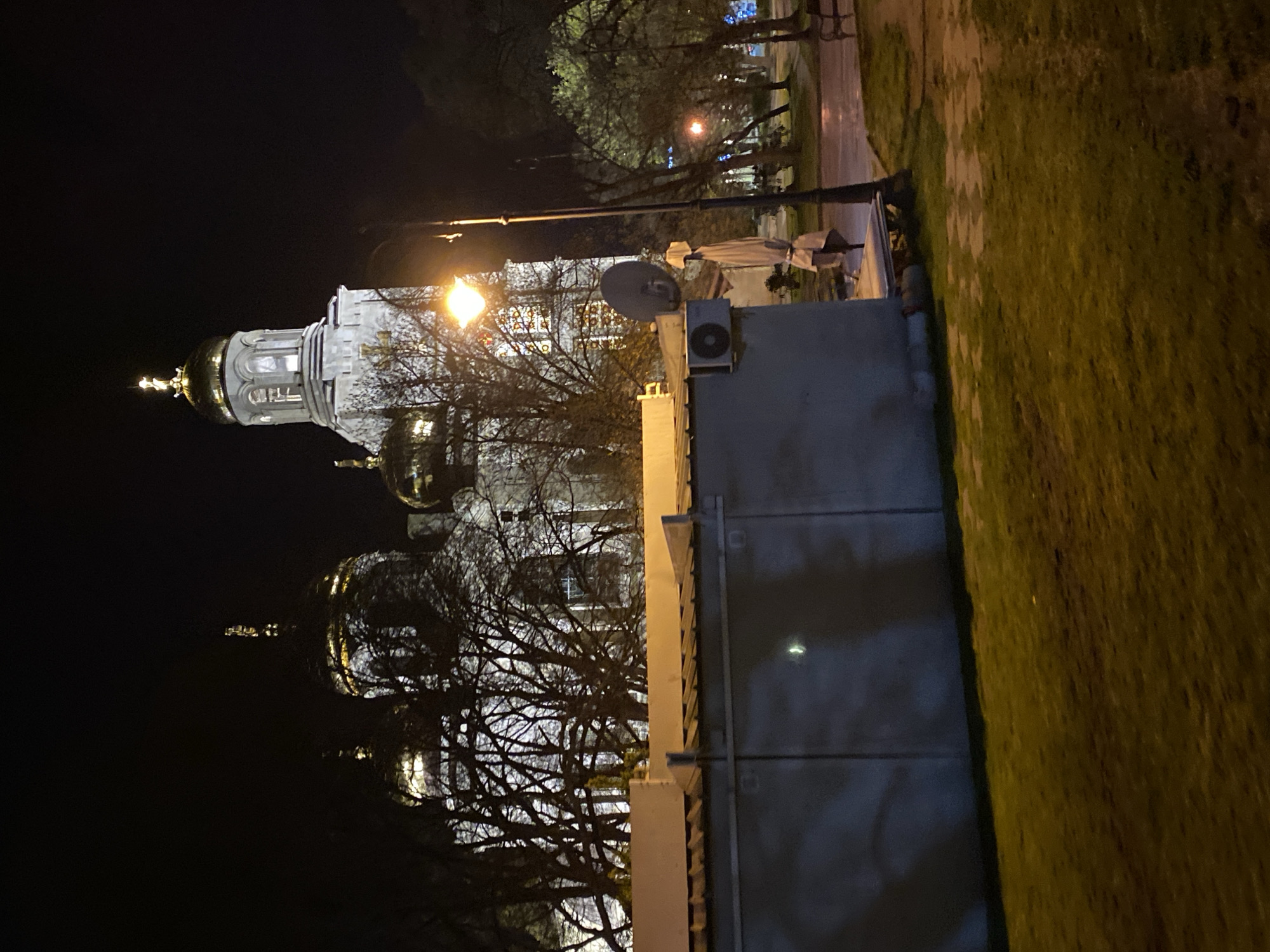 The image shows the Dormition of the Mother of God Cathedral in Varna, Bulgaria at night. The cathedral's domes are illuminated, and trees surround it. A wall and grassy area are visible in the foreground. A lamppost stands near the wall. A satellite dish and air conditioning unit are mounted on a nearby building. The lighting indicates it is nighttime. The street is wet with possible rain. Other distant lights indicate buildings and street lighting.