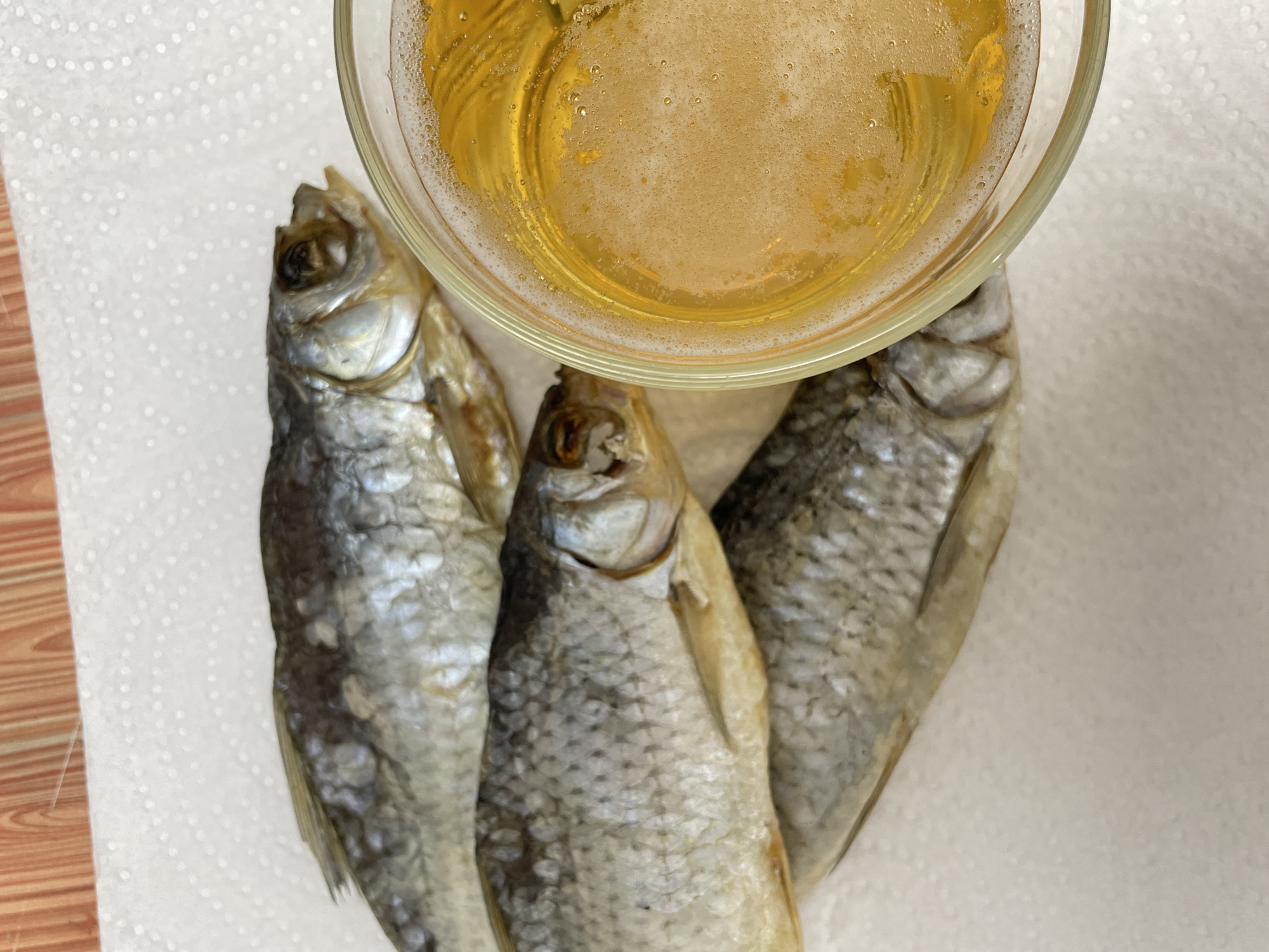Three dried fish lie beside a glass of beer on a white napkin. The scene evokes the common pairing of dried fish and beer, often enjoyed as a snack or appetizer. The backdrop lacks specific details that would pinpoint the location to Izhevsk, Russia, or any other place.
