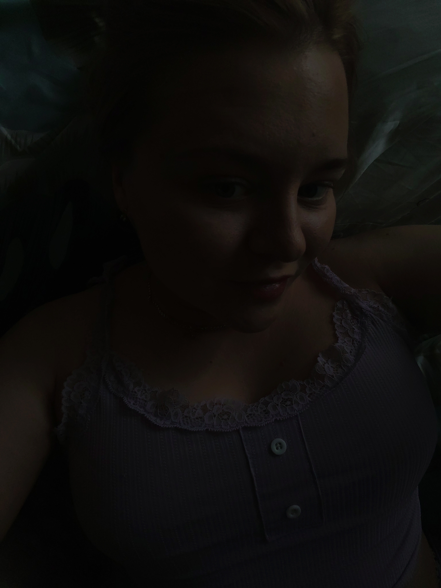 A person with light skin is lying down wearing a light purple tank top with lace trim and two buttons. The background appears to be a dark-colored fabric. The lighting is low, making it difficult to discern specific details. There is no evidence in the image to suggest a location in Izhevsk, Russia, or any other specific city.