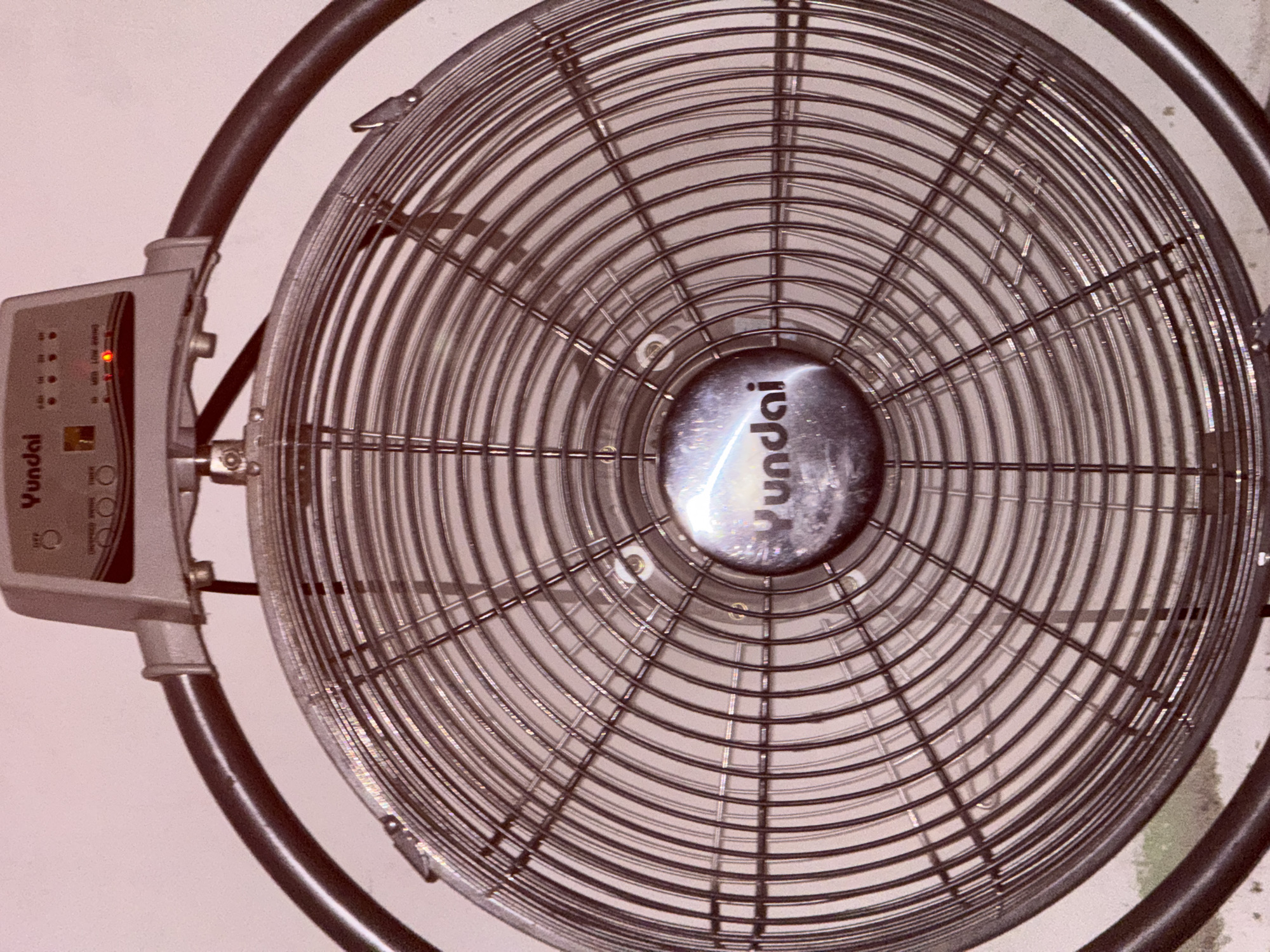The image shows a close-up, low-angle view of a metal oscillating fan. The fan has a circular grille with a dark outer rim. In the center of the grille is a circular emblem with the brand name "Yundai" written on it. Above the fan, a control panel is visible with buttons and indicator lights. One light, labeled "SWING," is illuminated in red. The fan is positioned indoors, against a light-colored wall. There is no information or context provided in the image to indicate the location in Babahjurong, Indonesia. The scene is static and shows no people or actions.