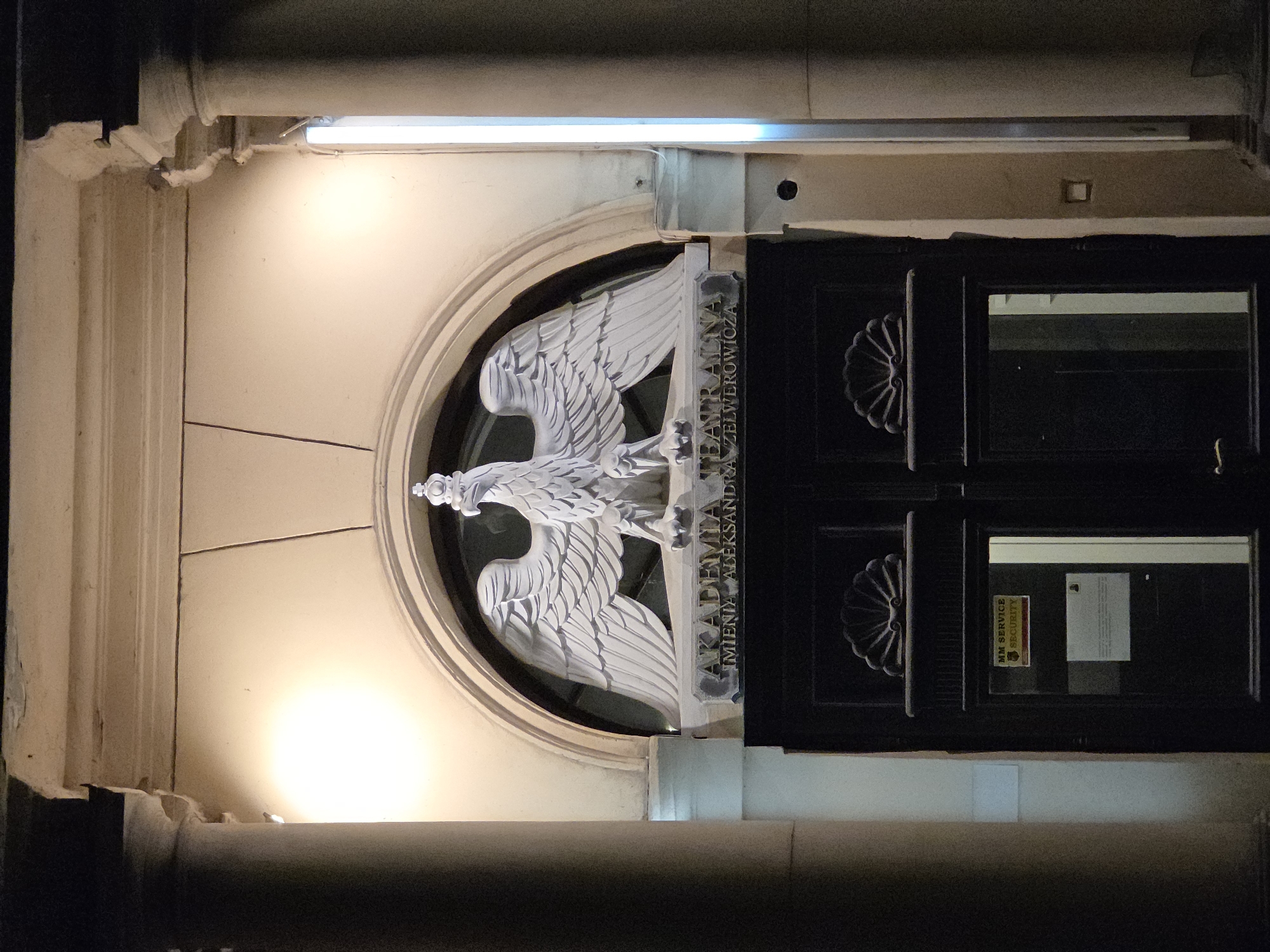 The image shows the entrance to the Aleksander Zelwerowicz National Academy of Dramatic Art in Warsaw, Poland. Above the black wooden double doors is a white eagle sculpture mounted within a circular recess in the building's facade. The eagle is holding a plaque bearing the name "AKADEMIA TEATRALNA IMIENIA ALEKSANDRA ZELWEROWICZA." The building's facade is off-white, and the entrance is illuminated by a light fixture above. A "MM SERVICE SECURITY" sign is visible on the inside of the door, partially obscured by the glass. The building appears to be constructed of stone, as evidenced by the columns on either side of the entrance. The surrounding environment is dark, suggesting the photo was taken at night.