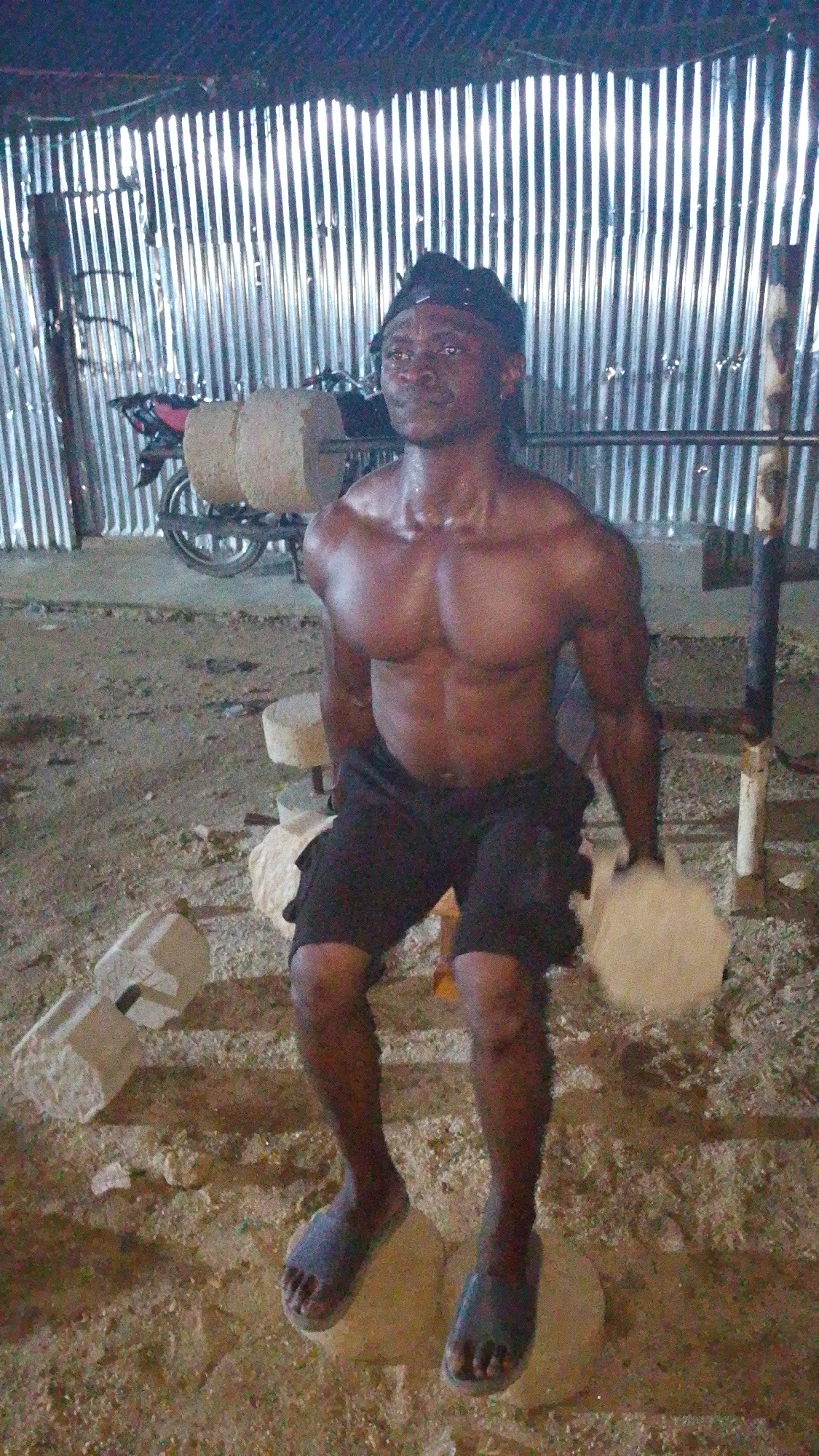 The image depicts a muscular Black man wearing a black cap and black shorts, sitting on large stones in what appears to be an outdoor gym or training area. The background consists of a corrugated metal wall and some exercise equipment, including what looks like a weight rack and possibly a motorcycle in the distance. The ground is dirt and scattered with debris and more stones. The lighting suggests it is either dusk or nighttime. The man's body is glistening with sweat, indicating he has been exercising. The overall impression is of a rudimentary, self-made fitness facility. The location is identified as Indo, Nigeria.