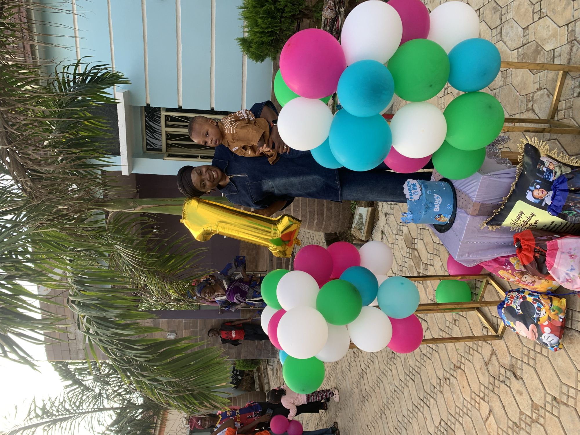 The scene depicts an outdoor birthday celebration, consistent with a residential area in Jos, Nigeria. In the foreground, two decorative balloon pillars, composed of white, green, magenta, and teal balloons, flank a small table. The table is draped with a sparkly light purple fabric and holds a blue two-tier birthday cake featuring "FAHEEM @ ONE" in white icing, a crown topper, and "Bluey" characters. On the ground adjacent to the table are several gift or party favor bags, including one with Mickey Mouse characters, another with Disney princesses, and a pink floral drawstring bag. A black decorative pillow or frame with "Happy Birthday Son" text and two small children's photos is also present.
At the center, a dark-skinned woman with a headwrap, wearing a dark denim-like long-sleeved top and matching wide-leg trousers, smiles towards the viewer. She holds a large gold foil balloon shaped like the number "1" in her left hand and a dark-skinned baby in a light brown patterned shirt and pants in her right arm. The baby looks slightly to its right.
To the left, several individuals are visible: a person partially obscured wearing a "Unicef Barcelona" jersey, a small child facing away holding a pink balloon, and a teenager in a red beanie and dark top further back. To the right and behind the central woman, a male teenager in a red t-shirt and black trousers stands, looking left. Beside him, a dark-skinned woman in a traditional patterned wrapper dress carries a baby on her back.
The ground is paved with light-colored, interlocking concrete pavers. The background features several large palm trees, partially obscuring a light-colored building with brick accents on the left and a light blue painted building with white horizontal trim on the right. A dark green shrub is visible near the base of the blue building. The setting is brightly lit by daylight, indicating an outdoor daytime event.