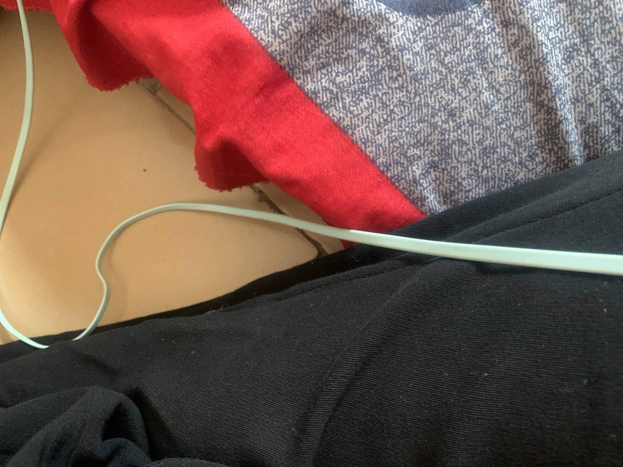 A light blue coat hanger is positioned diagonally across a scene of folded clothing. To the left is a dark fabric, likely pants or a skirt, with visible vertical seams. To the right, a bright red fabric is folded over a patterned fabric with a gray and blue design. The background appears to be a tiled surface. The image is an interior shot, and its specific location in Jauro Yinu, Nigeria, cannot be determined from the visual information provided.