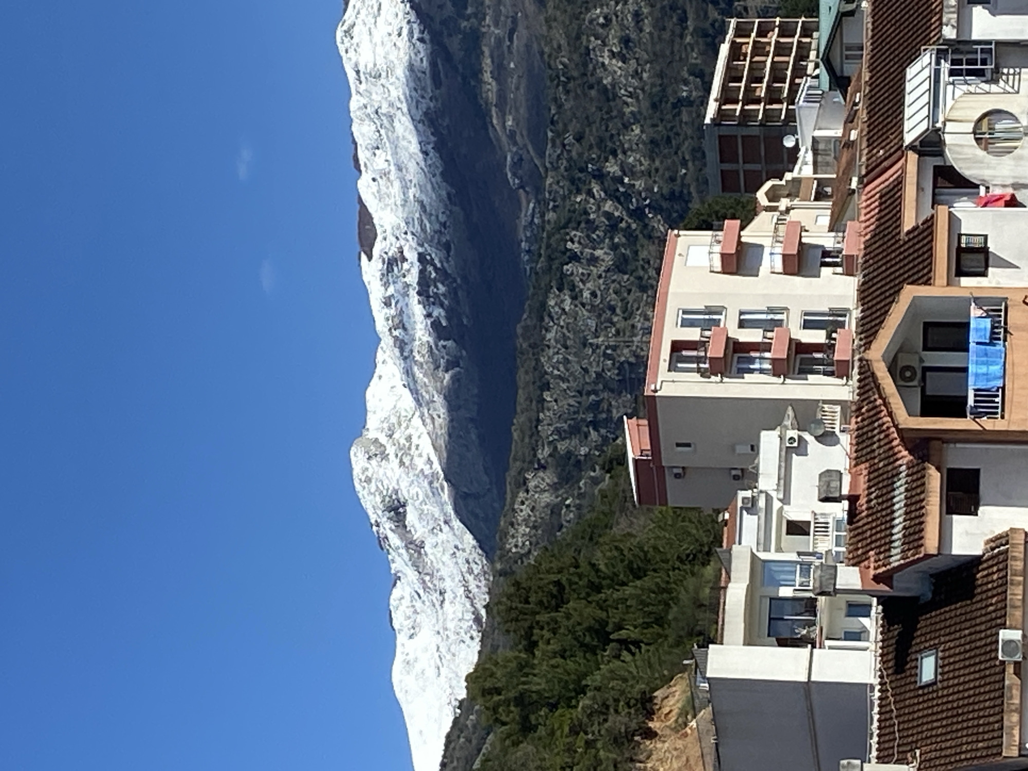 The image depicts a view in Budva, Montenegro, featuring a snow-covered mountain range to the left, a clear blue sky, lush green vegetation at the base of the mountains, and residential buildings with terracotta roofs and white facades in the foreground. Some buildings have visible air conditioning units and balconies. There are no people visible in the image. The mountain range dominates the left side of the frame, contrasting with the densely packed urban environment on the right.
