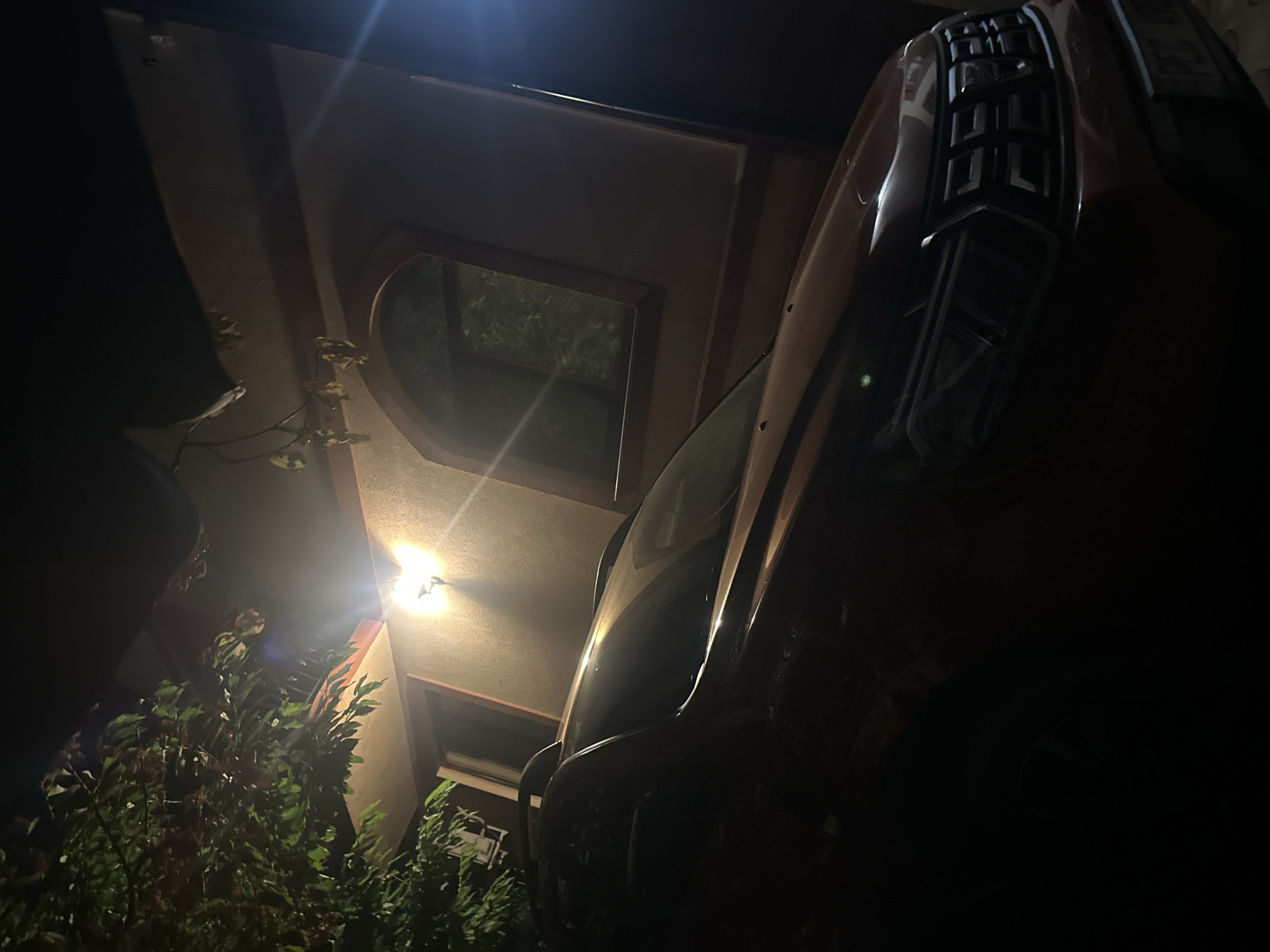 In Orhei, Moldova, a red car, possibly a Dacia Duster based on the front grille and headlight design, is parked at night in front of a building. The building features an arched window and an exterior light fixture that illuminates the scene. Lush green foliage from a tree overhangs the left side of the car and the building's roof. The overall lighting suggests it is nighttime, with artificial light sources casting shadows and creating a dramatic atmosphere. The license plate of the car is partially visible, showing "PEC 0".