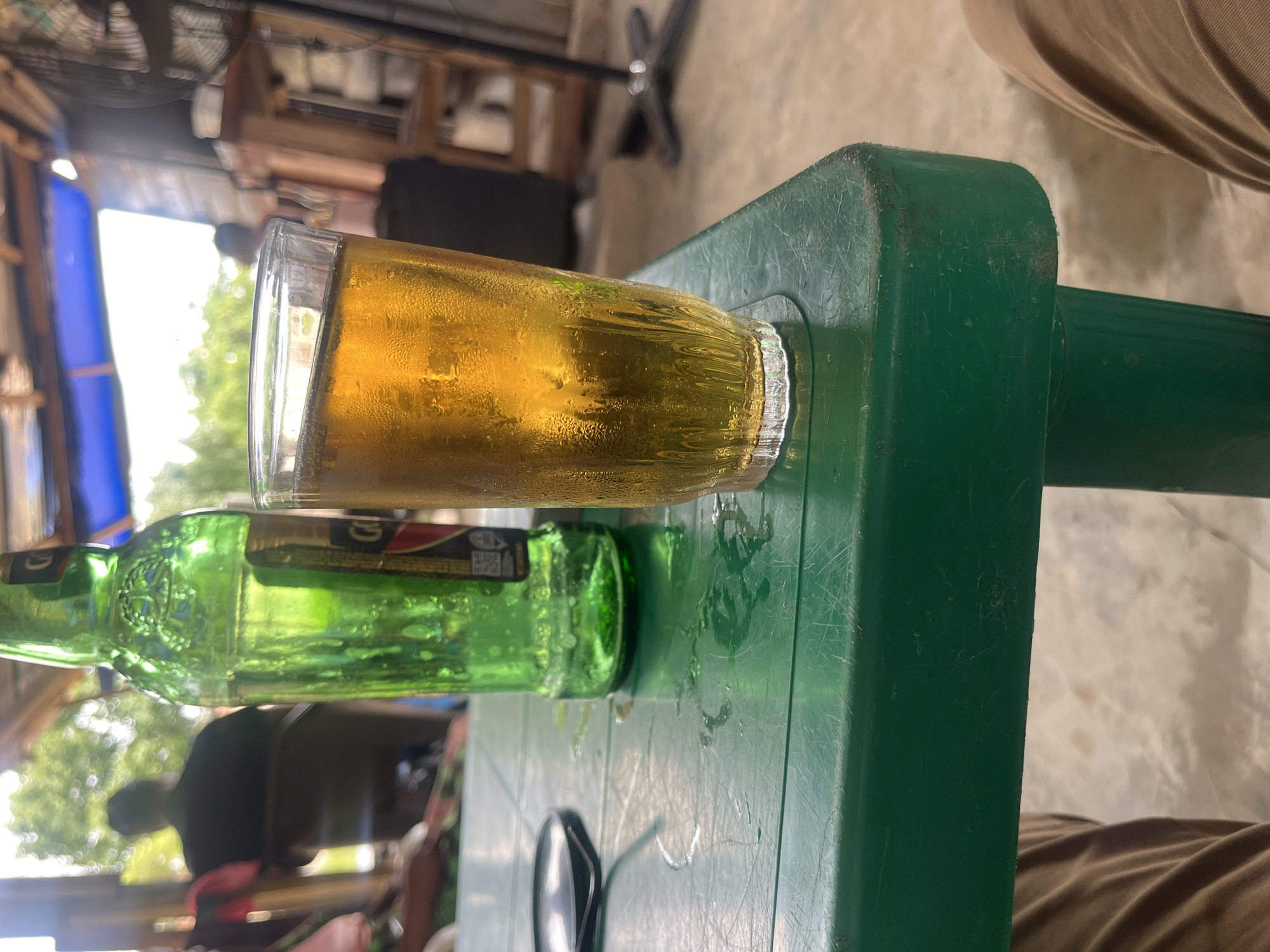 A green plastic table holds a glass of beer and a green beer bottle. There's condensation on the glass. Sunglasses are on the table as well. Part of a person's leg is visible next to the table. The background suggests an outdoor setting, possibly in Osogbo, Nigeria.