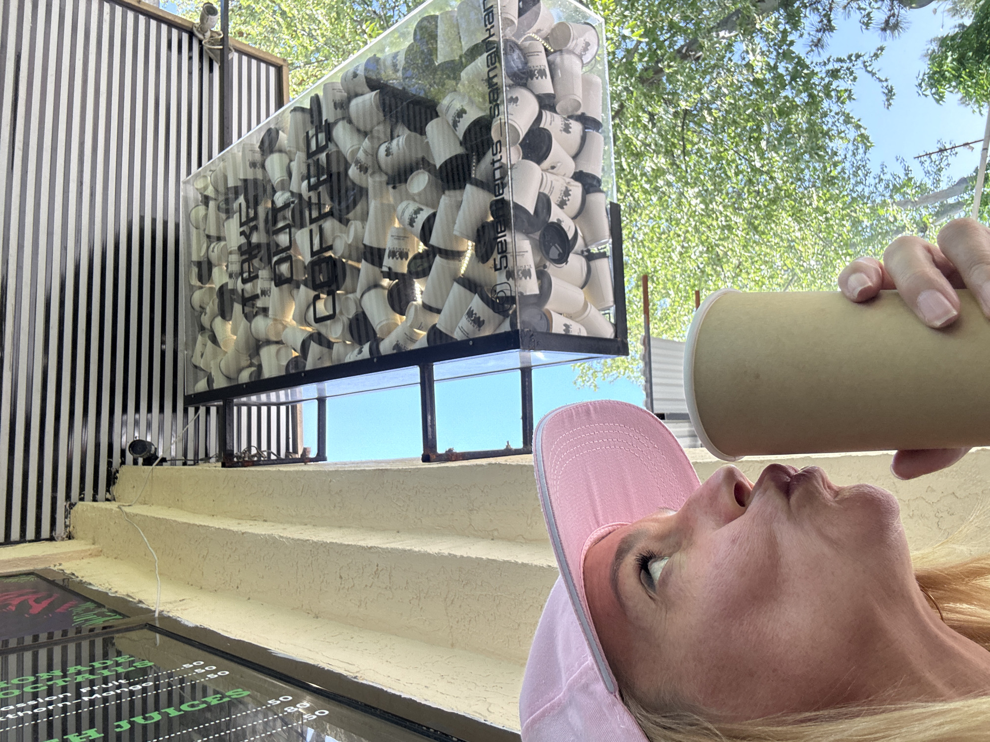A blond-haired, fair-skinned woman wearing a pink baseball cap is tilting her head back and drinking from a cylindrical, tan-colored cup. Her gaze is directed upwards. The scene is outdoors. Behind her is an elevated structure painted in a light, creamy color with textured stucco-like surfacing, resembling steps. A large, rectangular, transparent display case filled with stacks of paper cups is positioned above the steps. The words "OFFee ELEMENTS Se.marikan" are printed in a stylized font on the paper cups. To the left of the display case is a facade with vertical black and white stripes. At the bottom left of the image, a menu with various drink options is visible behind a glass pane. Overhead, green tree foliage and patches of blue sky are visible. The location is Samarkand City, Uzbekistan.